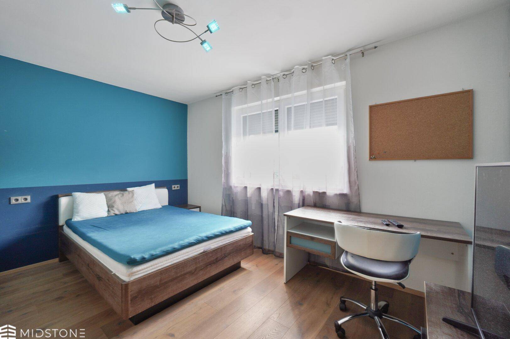 Schlafzimmer mit blau akzentuierter Wand, Holzbett und Schreibtisch am Fenster.
