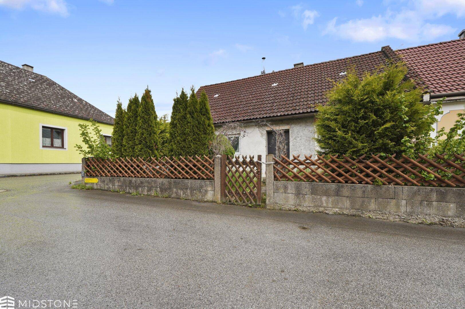 Vorderansicht des Hauses mit Zaun, Hecken und Bäumen entlang der Straße.