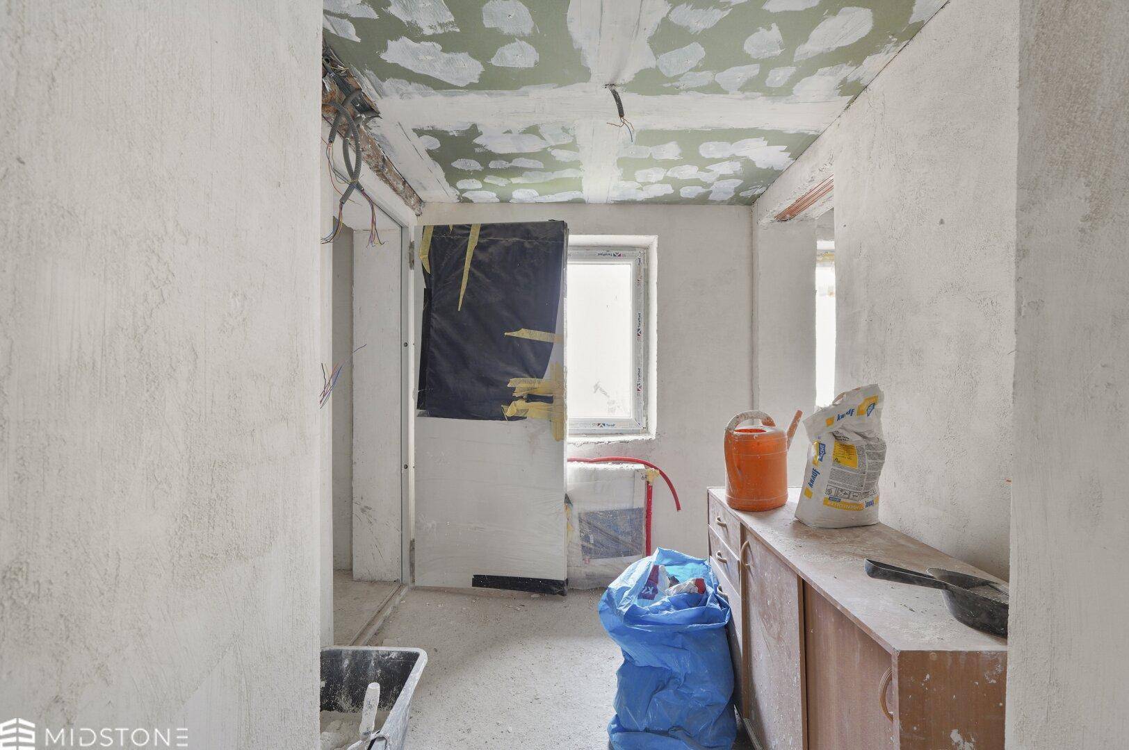Flur oder kleiner Raum im Rohbauzustand mit Fenster und Baumaterialien, bereit zur Fertigstellung.