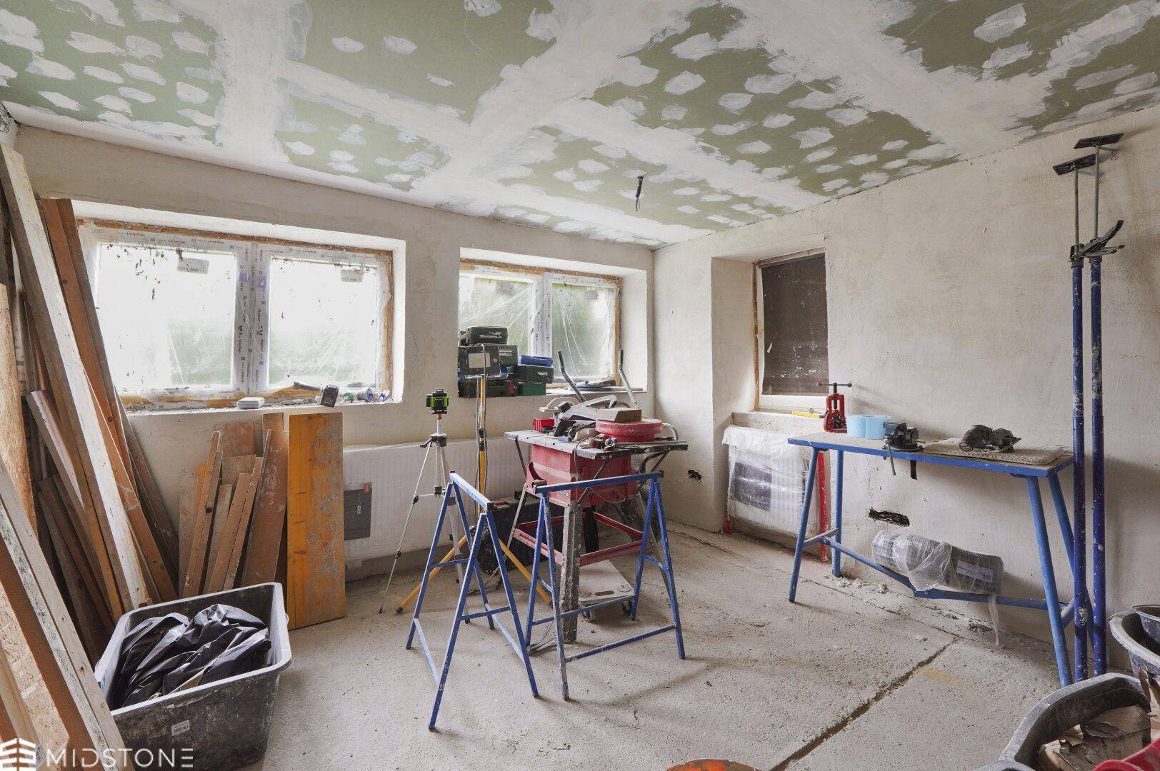 Renovierungsraum mit Baumaterialien und Werkzeugen, unfertigen Wänden und mehreren Fenstern.