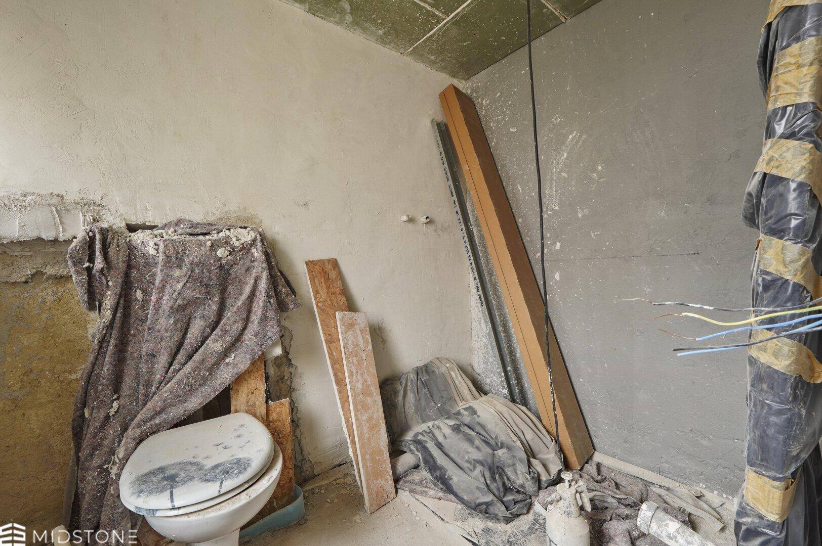 Badezimmer im Rohbau mit alter Toilette und Baumaterialien, bereit für die Sanierung.