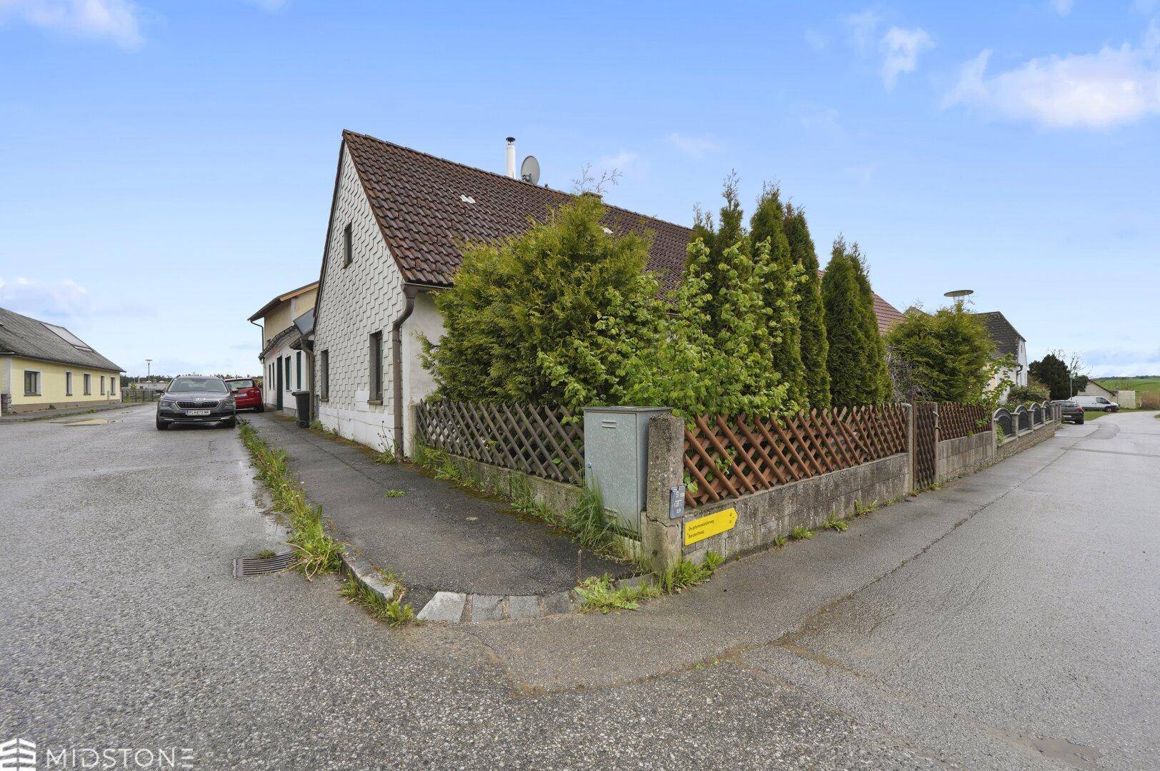 Eckansicht des Hauses mit Satteldach, Zaun und Vorgarten an einer ländlichen Straße.