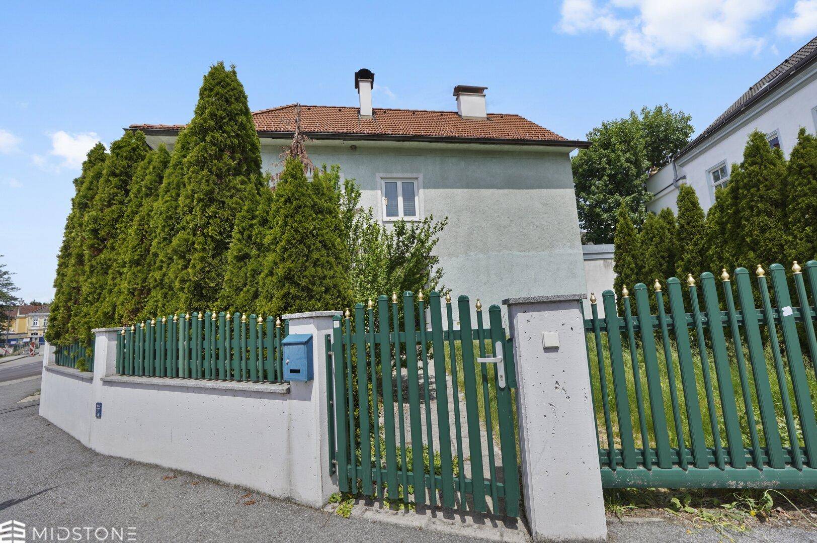 Seitenansicht eines Wohnhauses mit gepflegter Hecke und grünem Zaun unter blauem Himmel.