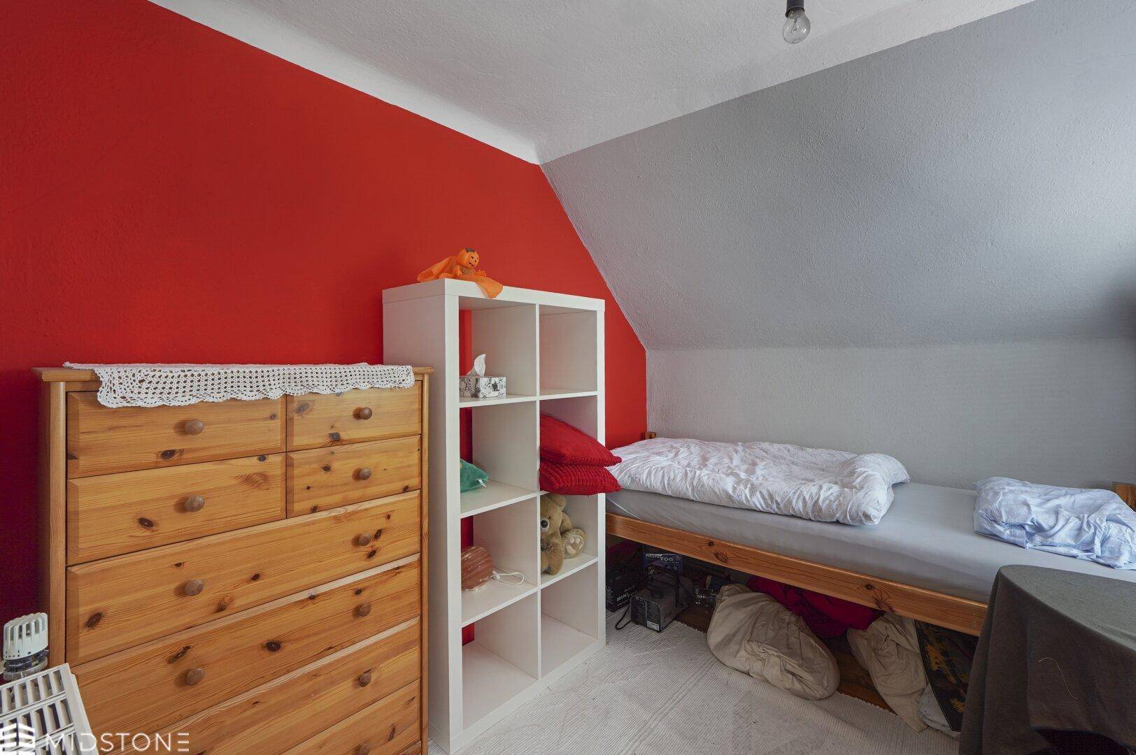 Schlafzimmer im Dachgeschoss mit roter Akzentwand, Einzelbett und Holzkommode unter schräger Decke.