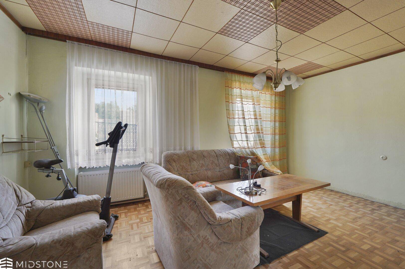 Wohnzimmer mit gemustertem Parkettboden, beiger Couch und einem Fenster mit Vorhängen.