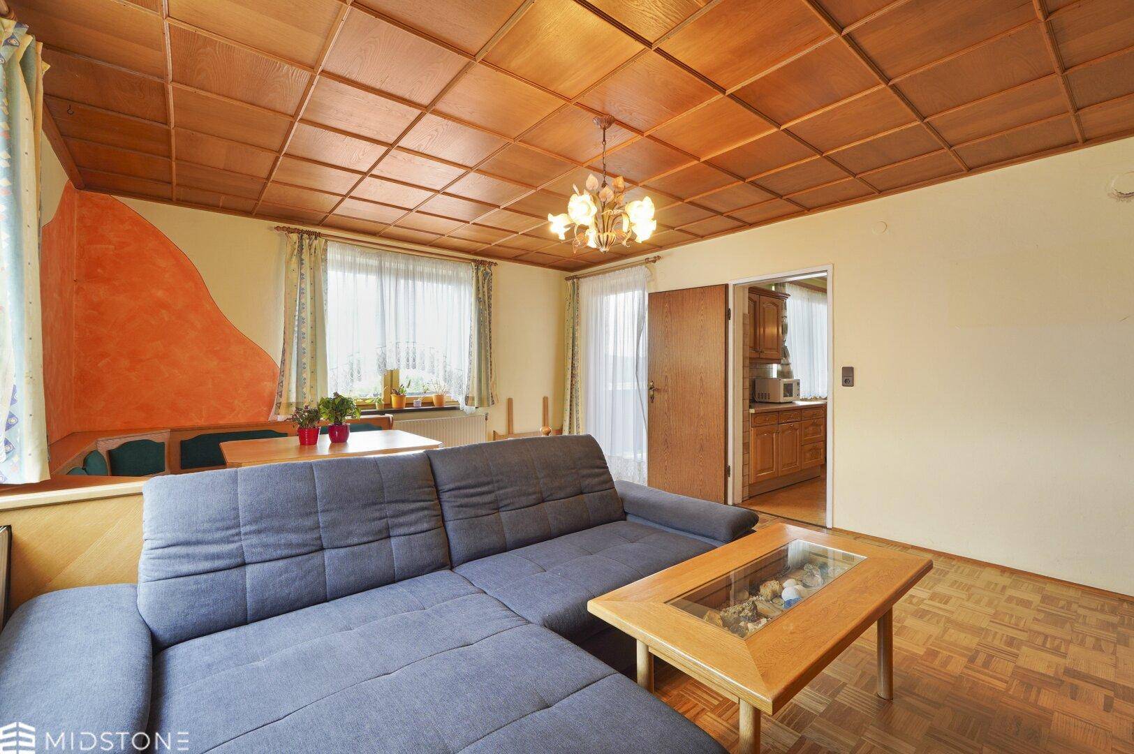 Helles Wohnzimmer mit Holzboden, Ecksofa und Zugang zur Küche.
