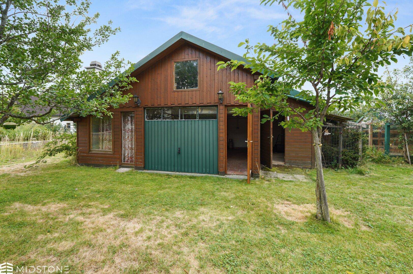 Rustikales Gartenhaus aus Holz mit grünen Toren und umgeben von Bäumen.