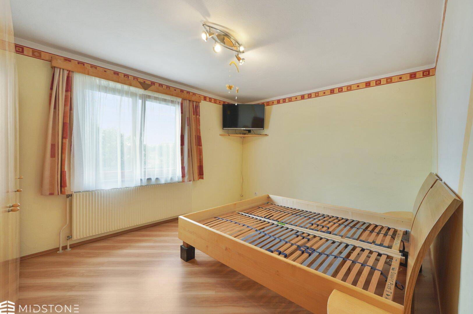 Helles Schlafzimmer mit Laminatboden, einem Bettgestell und Blick ins Grüne durch das Fenster.