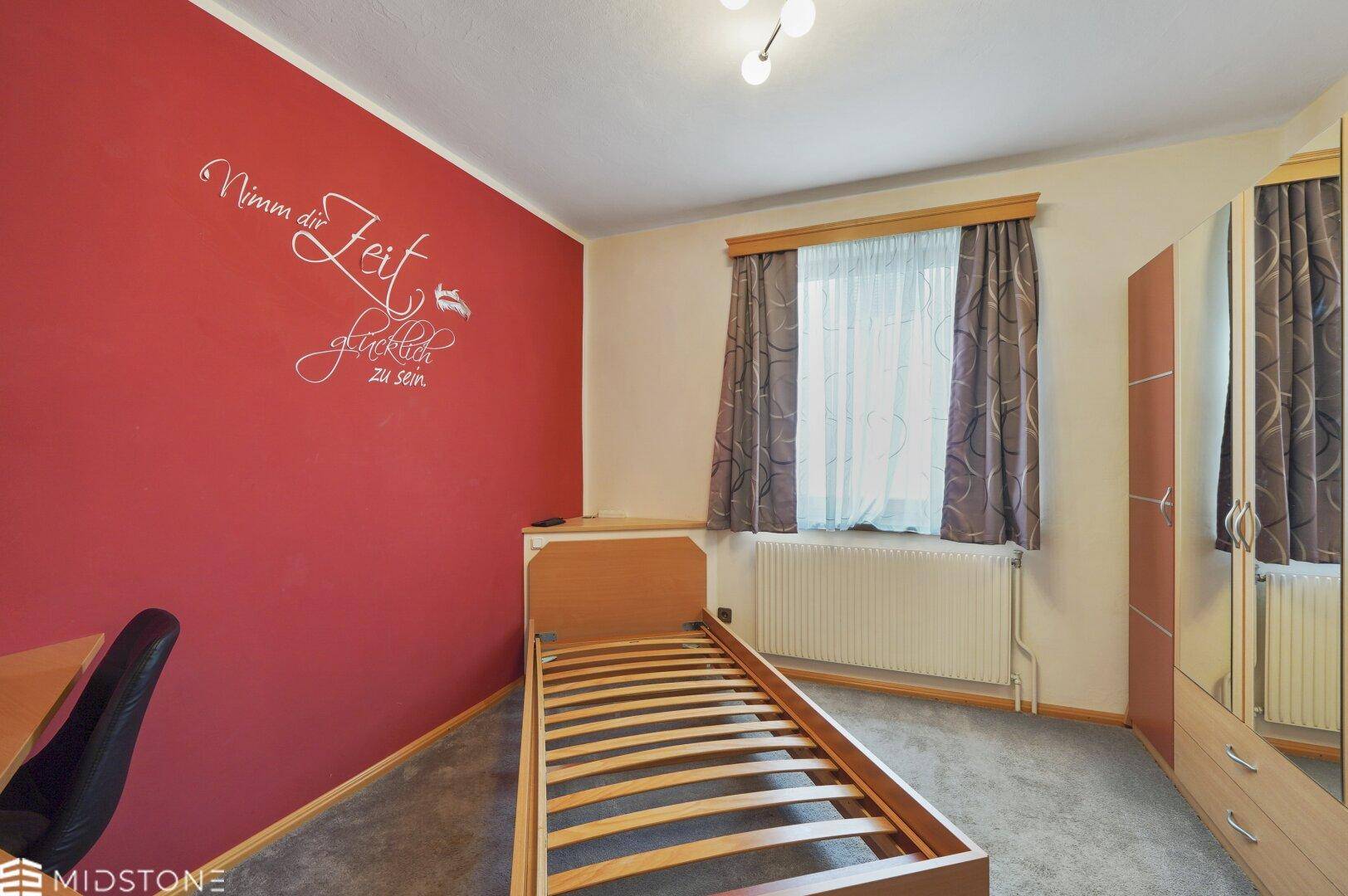 Gemütliches Schlafzimmer mit roter Akzentwand, Einzelbett und Teppichboden.