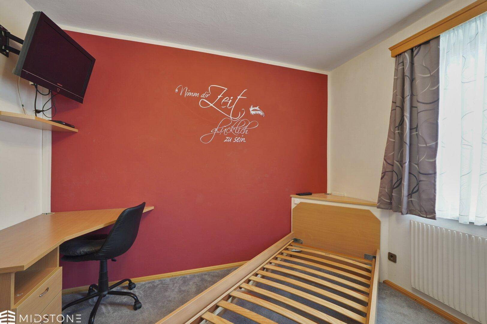 Schlafzimmer mit Schreibtisch, Einzelbett und roter Wandgestaltung.