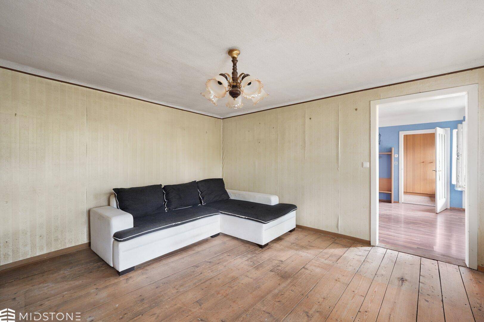Wohnzimmer mit Dielenboden und einem modernen Sofa, die Wände sind renovierungsbedürftig.