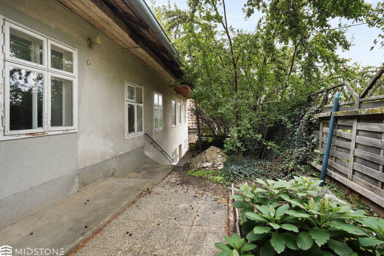 Seitenansicht des Hauses mit einem Weg, umgeben von üppigem Grün und einem Holzzaun.