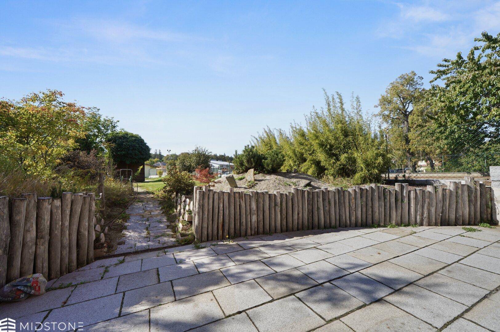 Gepflasterte Terrasse mit rustikalem Holzzaun und Zugang zu einem grünen Gartenbereich.