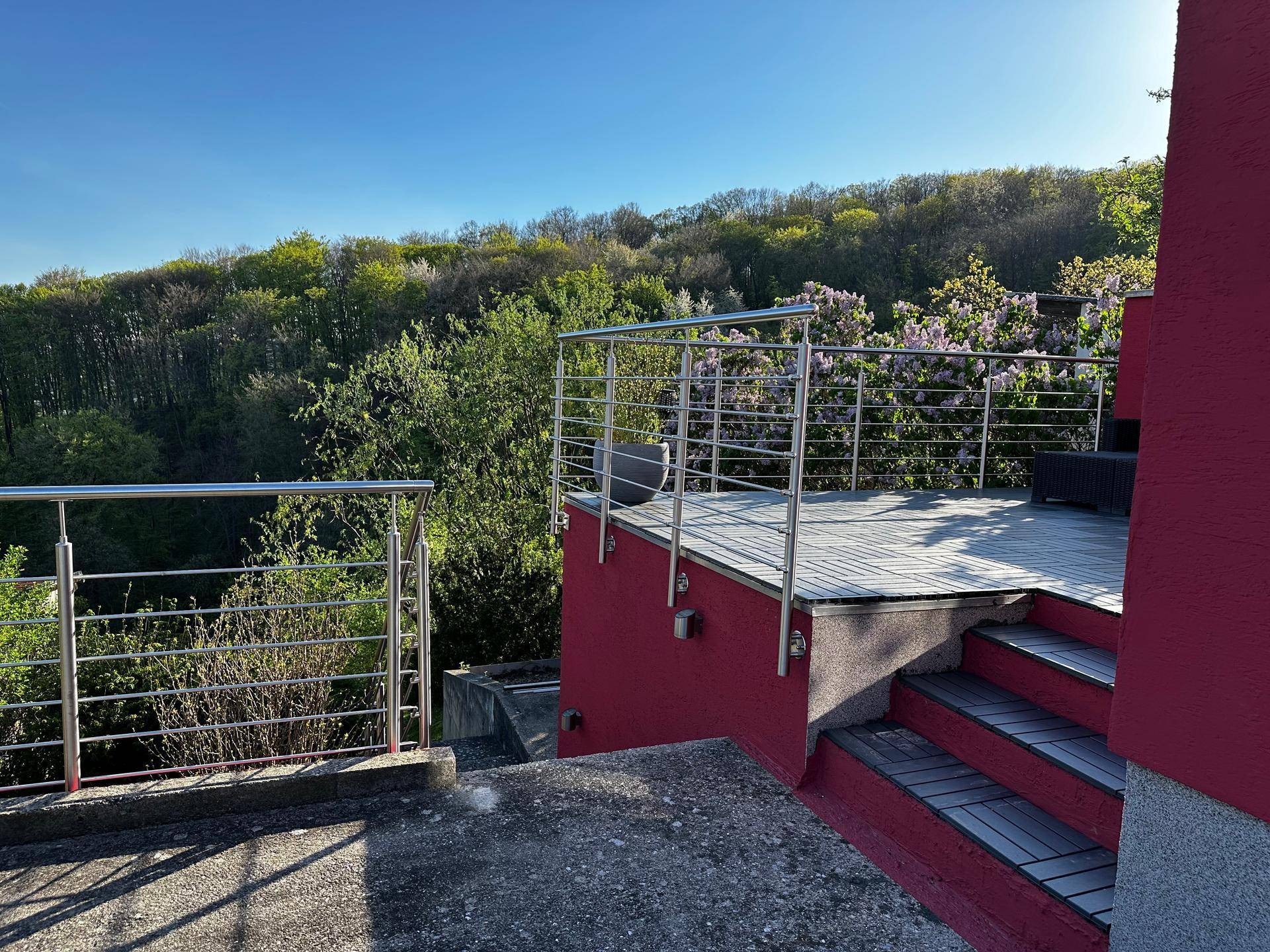 Terrasse mit Edelstahlgeländer und Treppenzugang, umgeben von üppiger Vegetation.