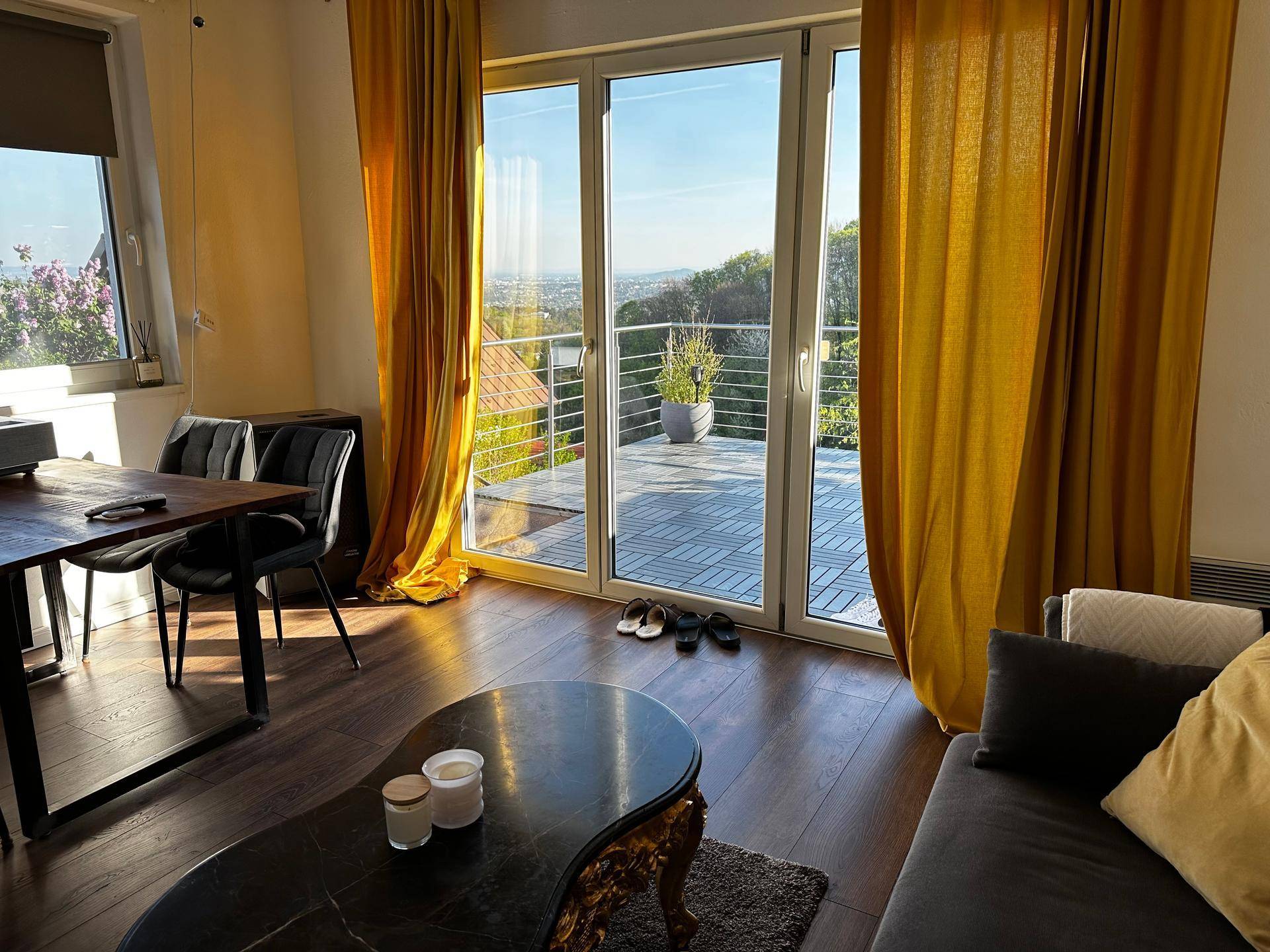 Helles Wohnzimmer mit Holzboden, gelben Vorhängen und Zugang zur Terrasse mit Weitblick.