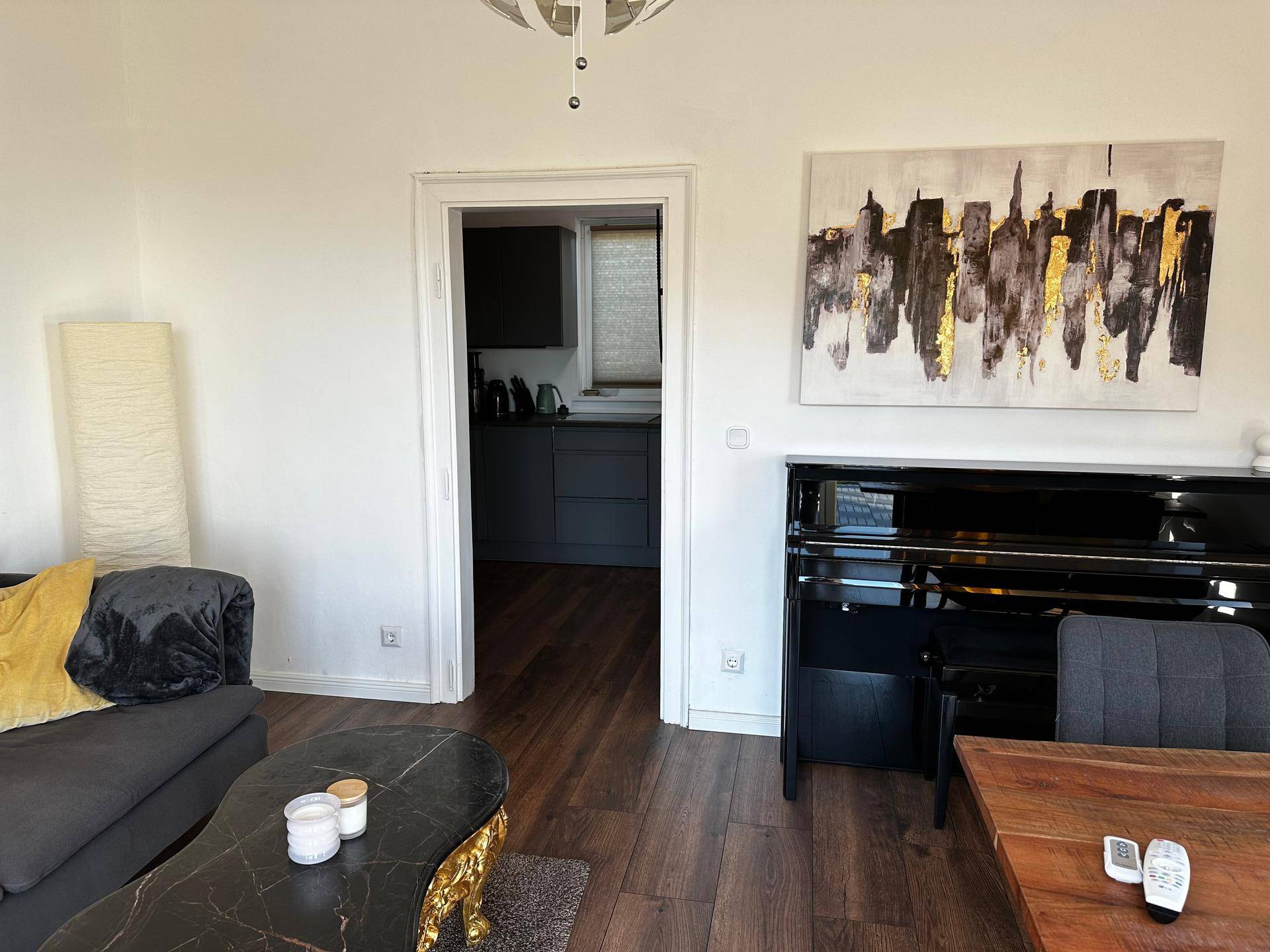 Gemütliches Wohnzimmer mit dunklem Holzboden, Klavier und Durchgang zur Küche.