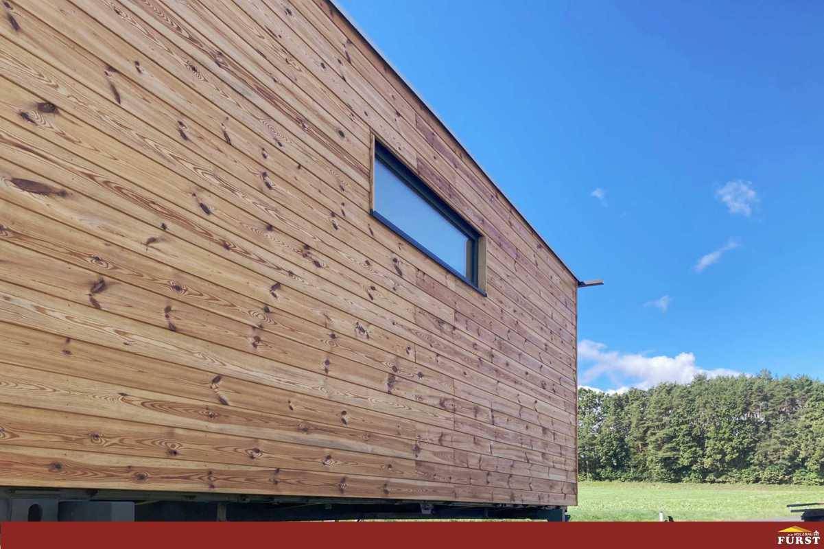 Seitenansicht des Tiny Houses mit Holzfassade und schmalem Fenster unter blauem Himmel.