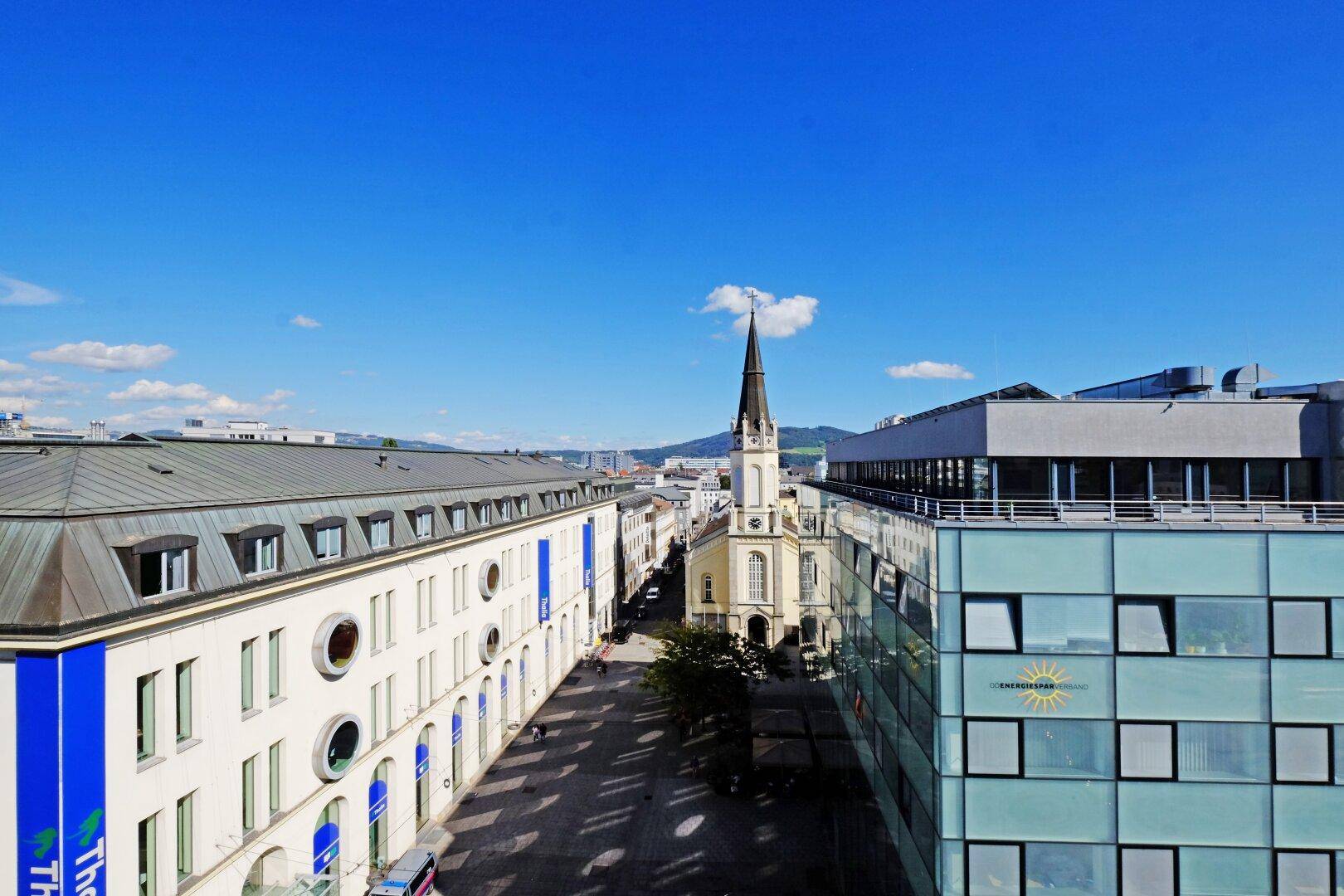 Panoramablick über eine Stadtstraße mit historischer Kirche und modernen Gebäuden unter klarem Himmel.