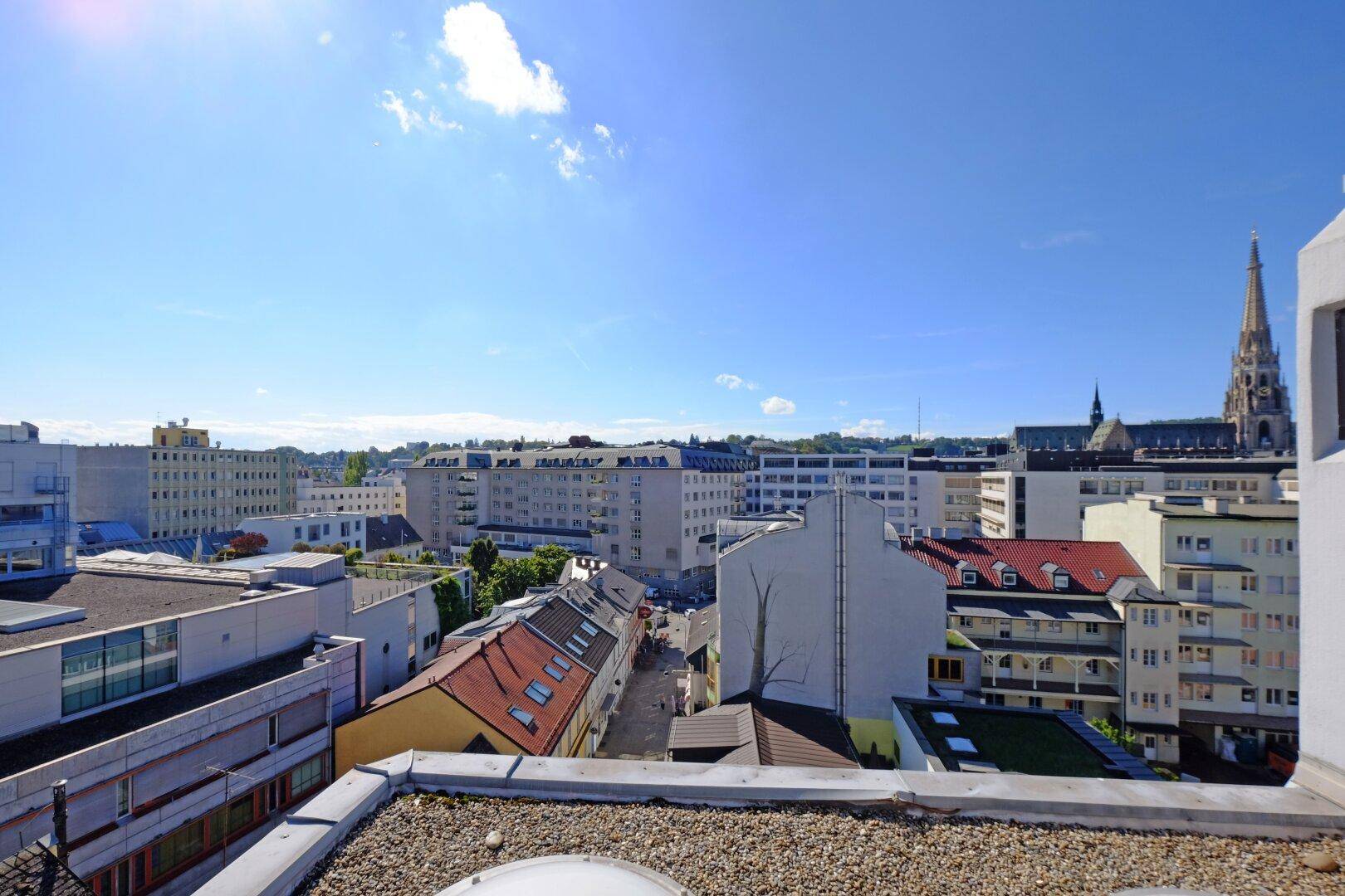 Blick von einem Dach über die Dächer der Stadt mit verschiedenen Gebäudetypen und einem Kirchturm in der Ferne.