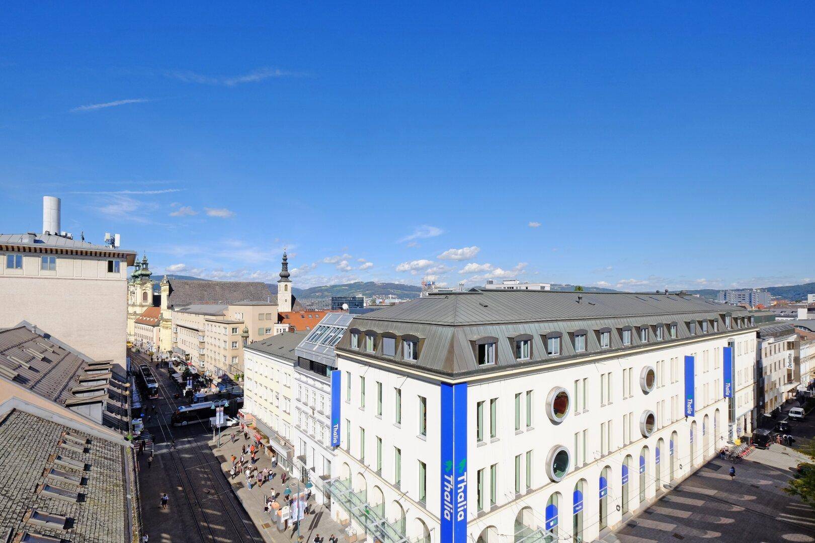 Panoramablick über eine belebte Stadtstraße mit einem großen Gebäude und Bergen im Hintergrund.