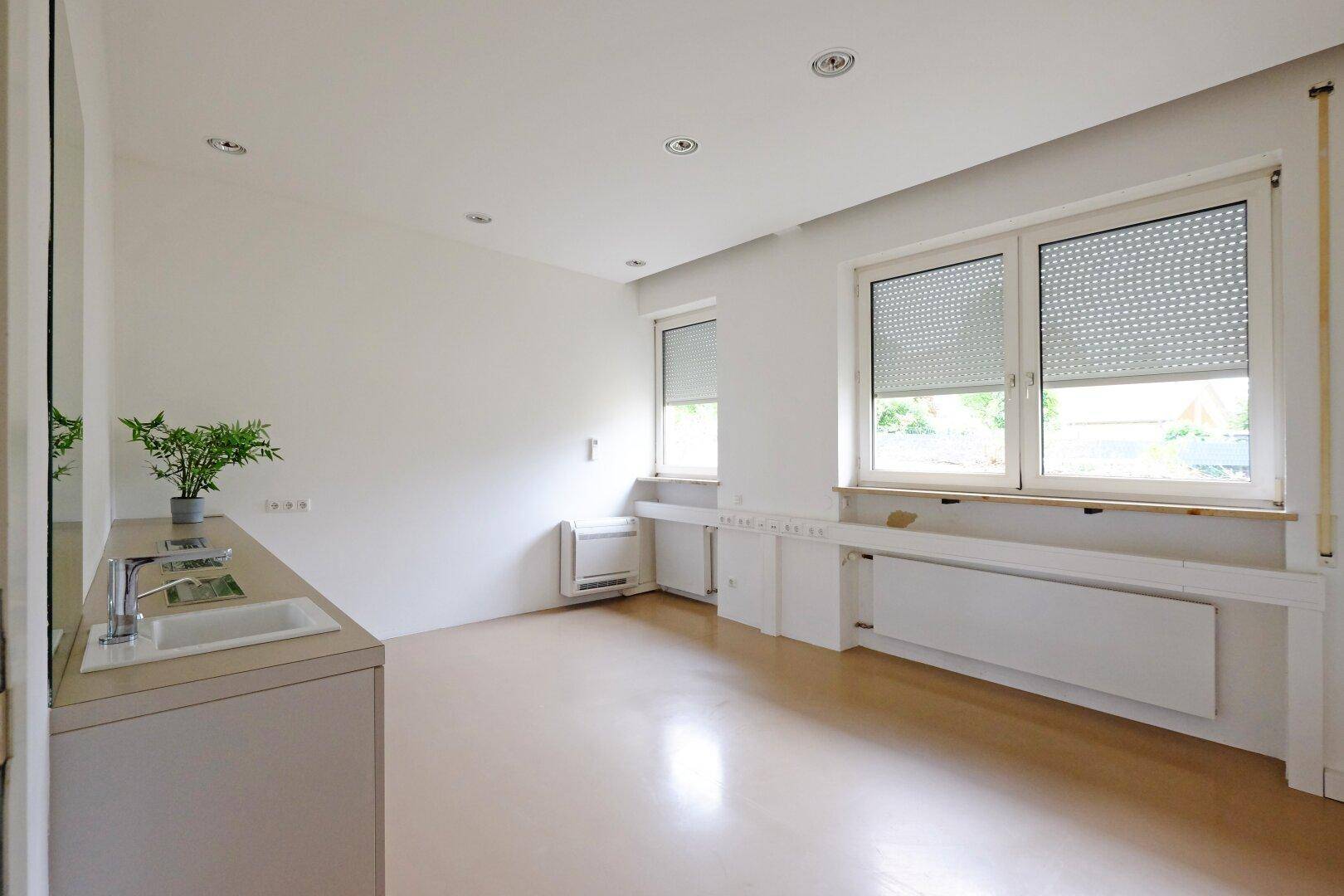 Helles Büro mit zwei Fenstern, Heizkörpern und einem Waschbeckenbereich.