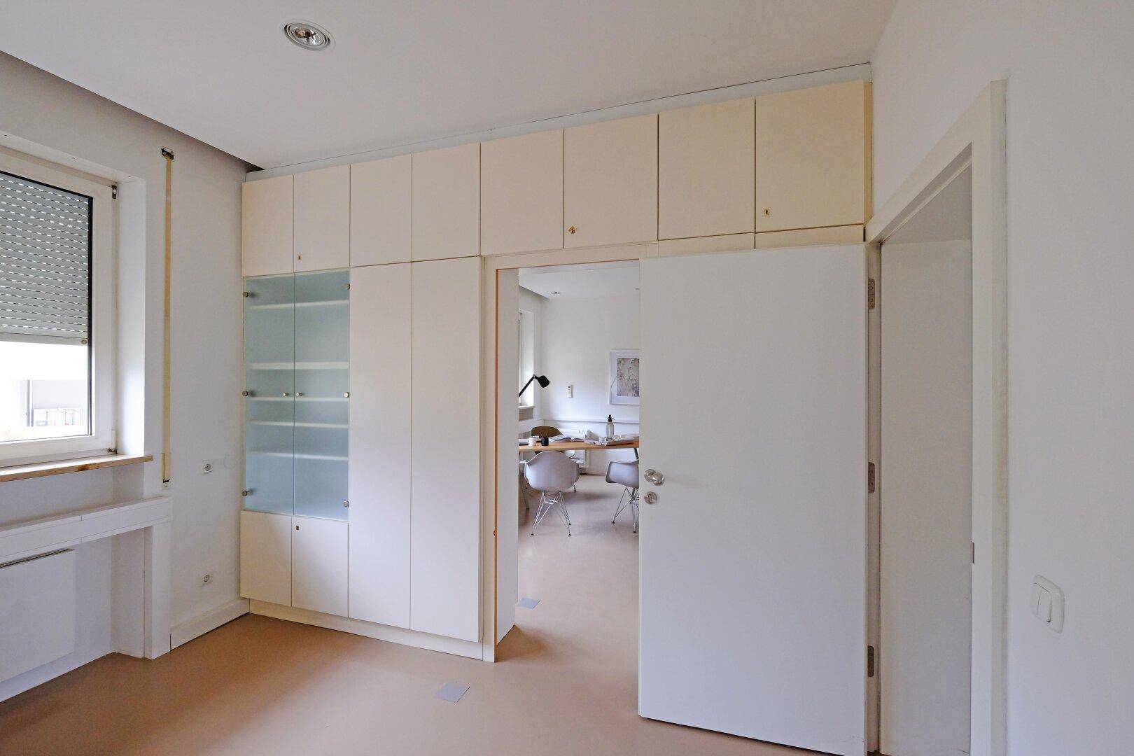Büro mit Einbauschränken, Glasvitrine und Zugang zu einem weiteren Arbeitsbereich.