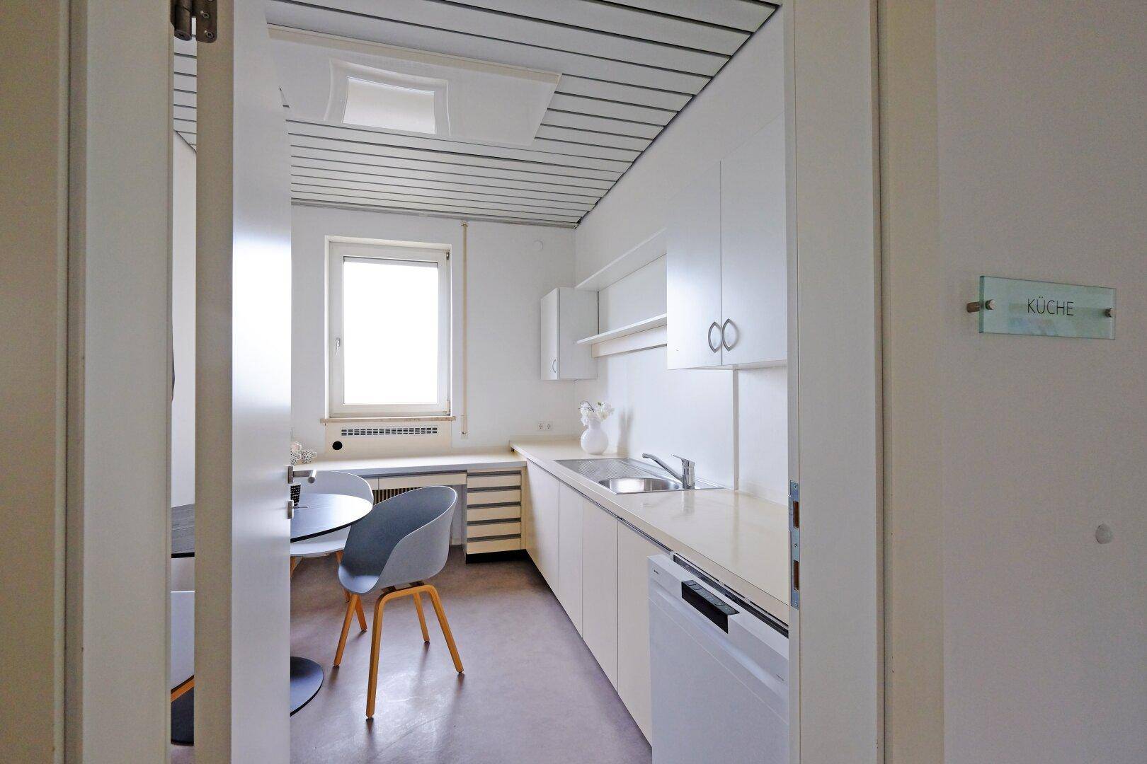 Funktionale Küche mit weißen Schränken, Spüle, Esstisch und Fenster.