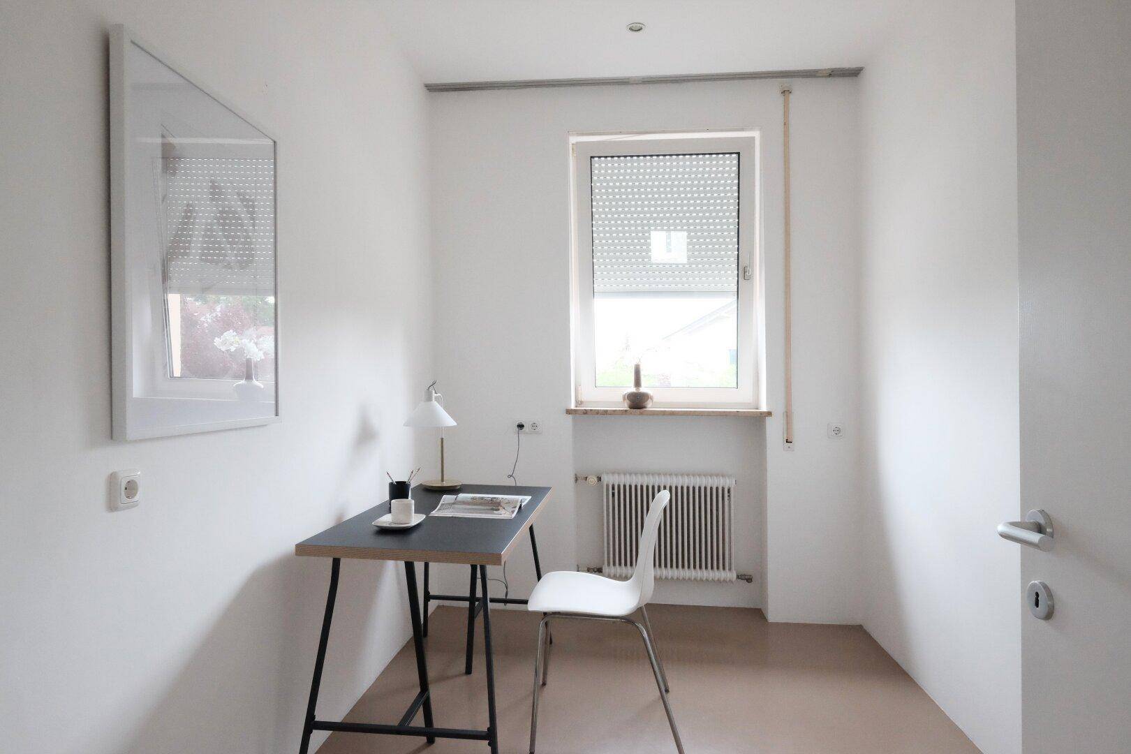 Kleines Büro mit Schreibtisch, Stuhl und Fenster mit Rollladen.