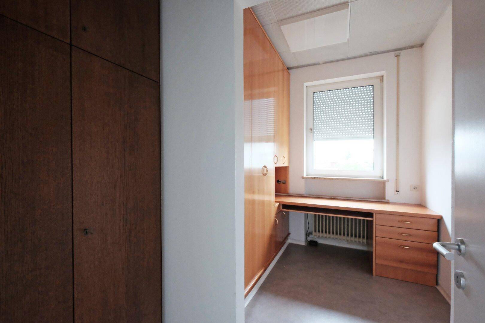 Büro mit Einbauschränken aus Holz, Schreibtisch und Fenster mit Rollladen.