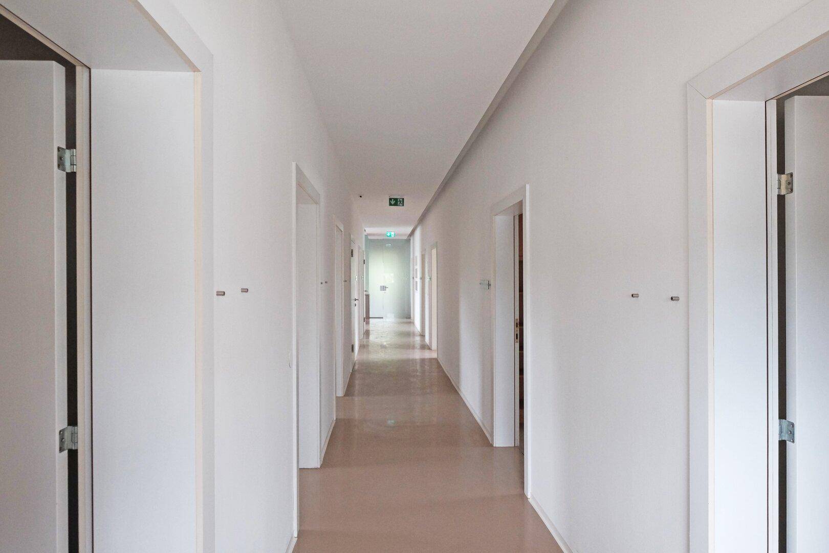 Langer, heller Flur mit mehreren Türen und Notausgangsbeschilderung.