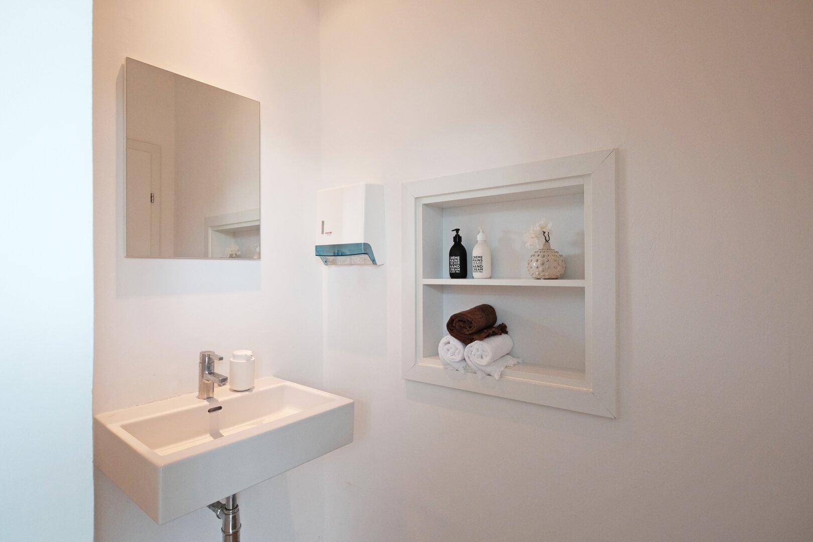 Modernes Badezimmer mit weißem Waschbecken, Spiegel und integriertem Regal für Handtücher und Pflegeprodukte.