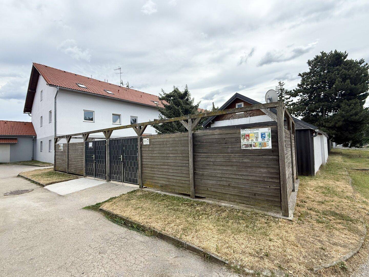 Außenbereich mit einer Holzumzäunung, wahrscheinlich für Mülltonnen, neben einem Wohngebäude.