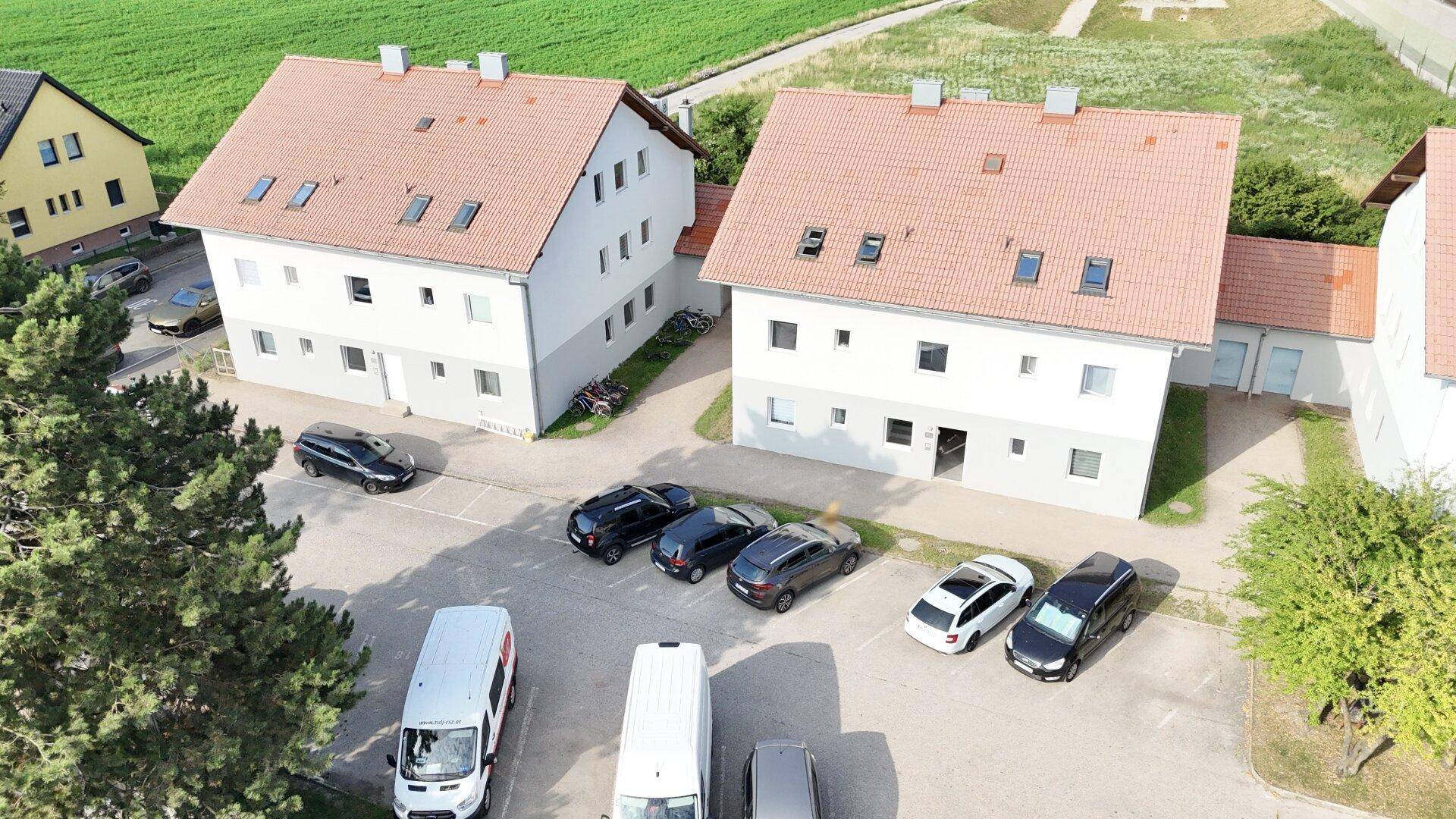 Luftaufnahme einer Wohnsiedlung mit mehreren Häusern und Parkplätzen unter blauem Himmel.