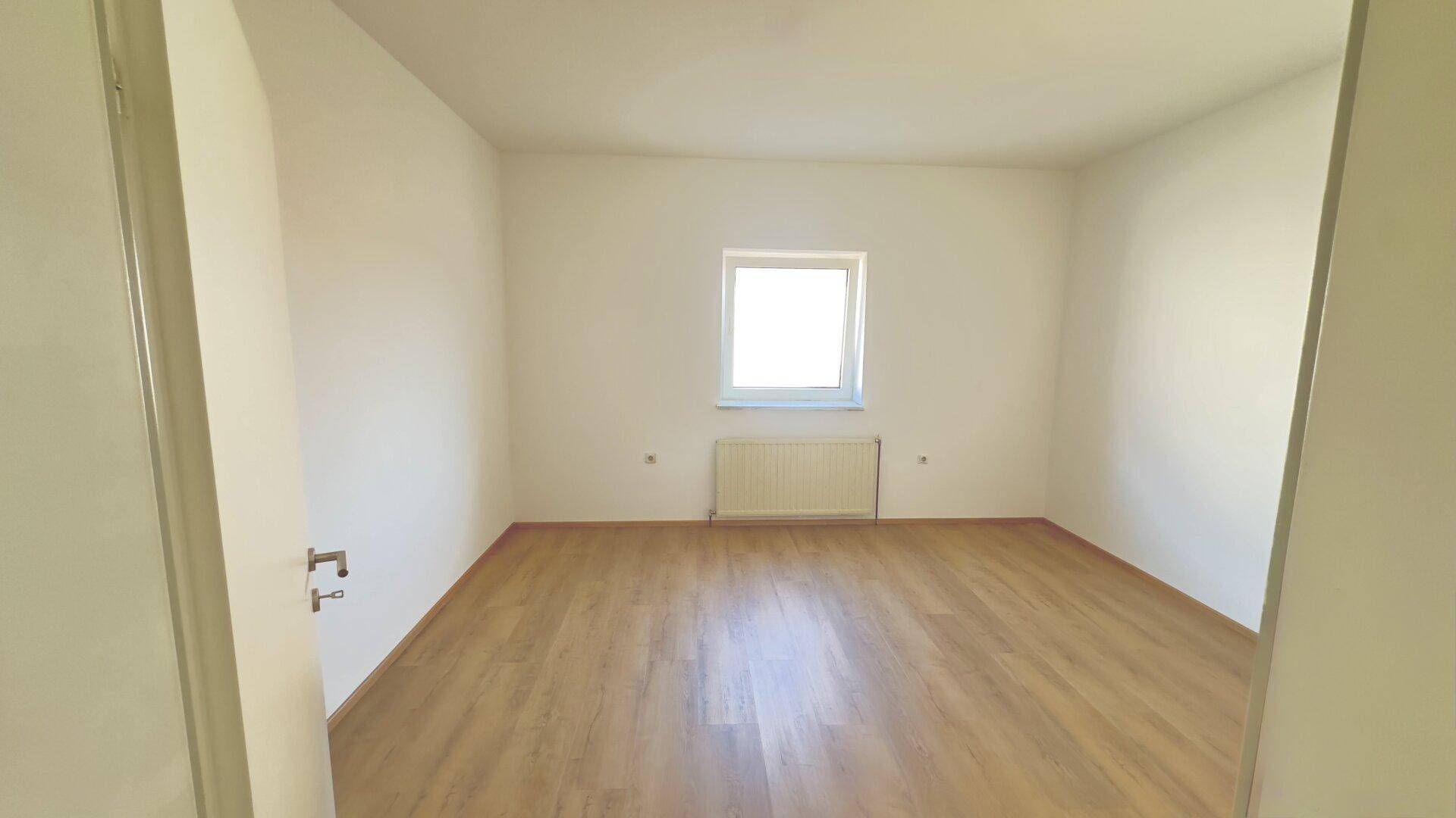 Leeres Zimmer mit hellem Laminatboden, weißer Wandfarbe und einem Fenster über dem Heizkörper.