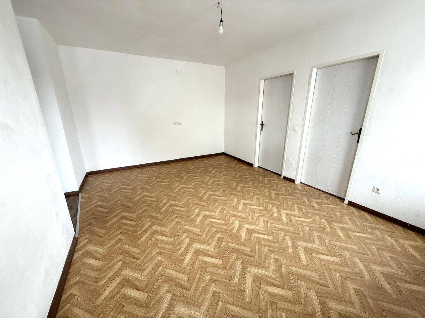 Leerer Raum mit hellem Fischgrät-Laminatboden und zwei weißen Innentüren.
