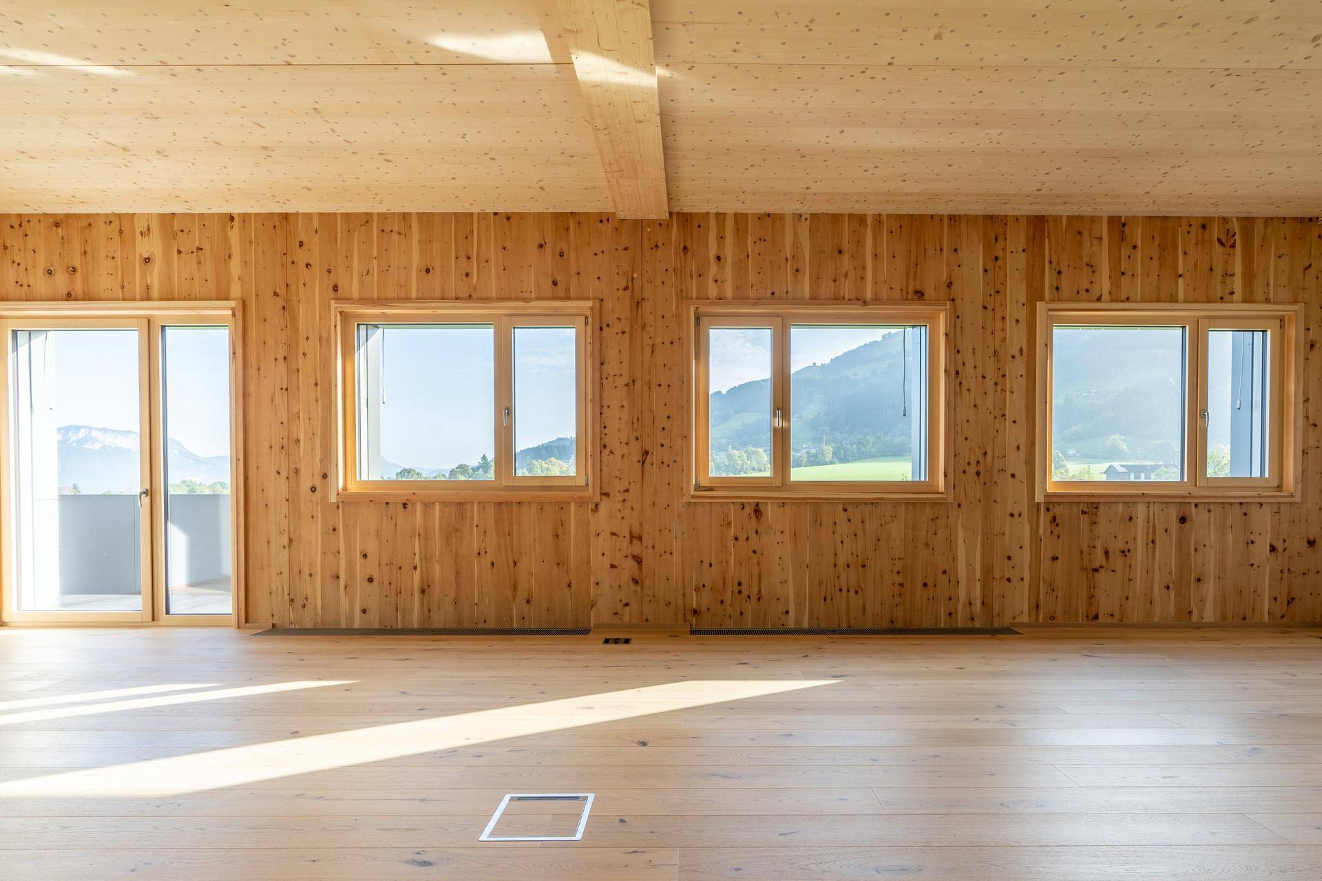 Großer, heller Raum mit Holzwänden und -decke, großen Fenstern und Zugang zu einem Balkon mit Bergblick.