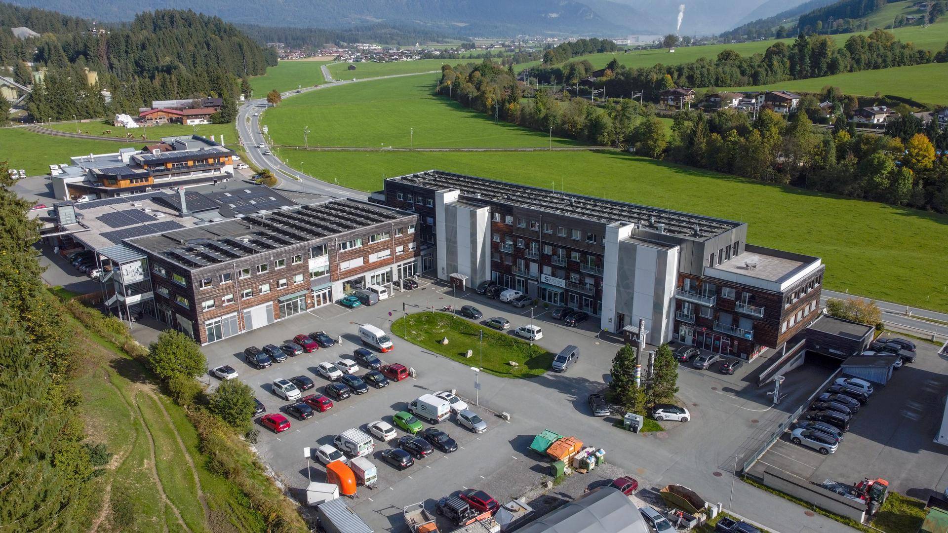 Luftaufnahme eines modernen Gebäudekomplexes mit Parkplätzen, umgeben von grünen Wiesen und Bergen.