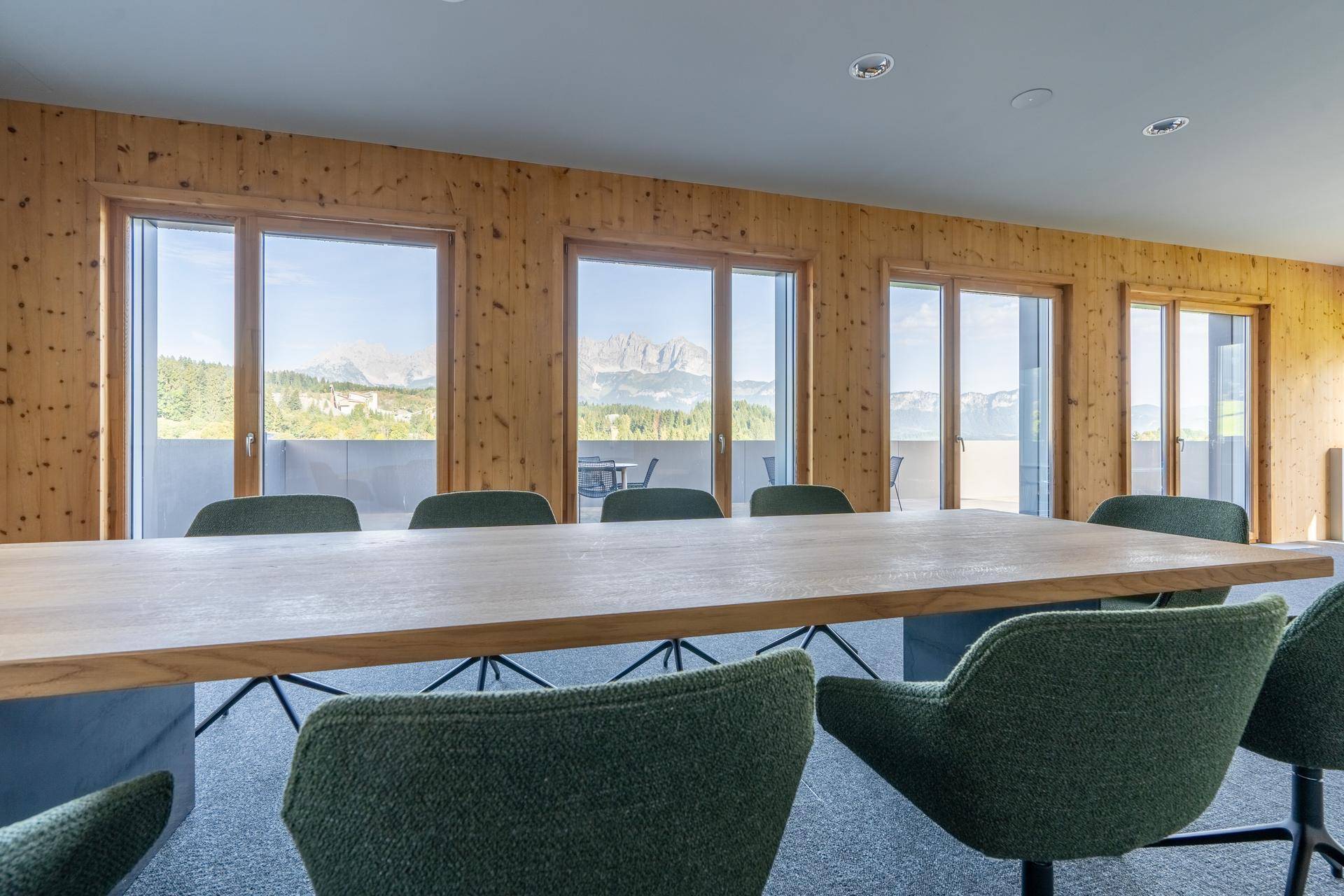 Konferenzraum mit langem Holztisch, grünen Stühlen und weitem Blick auf die Berglandschaft durch große Fenster.