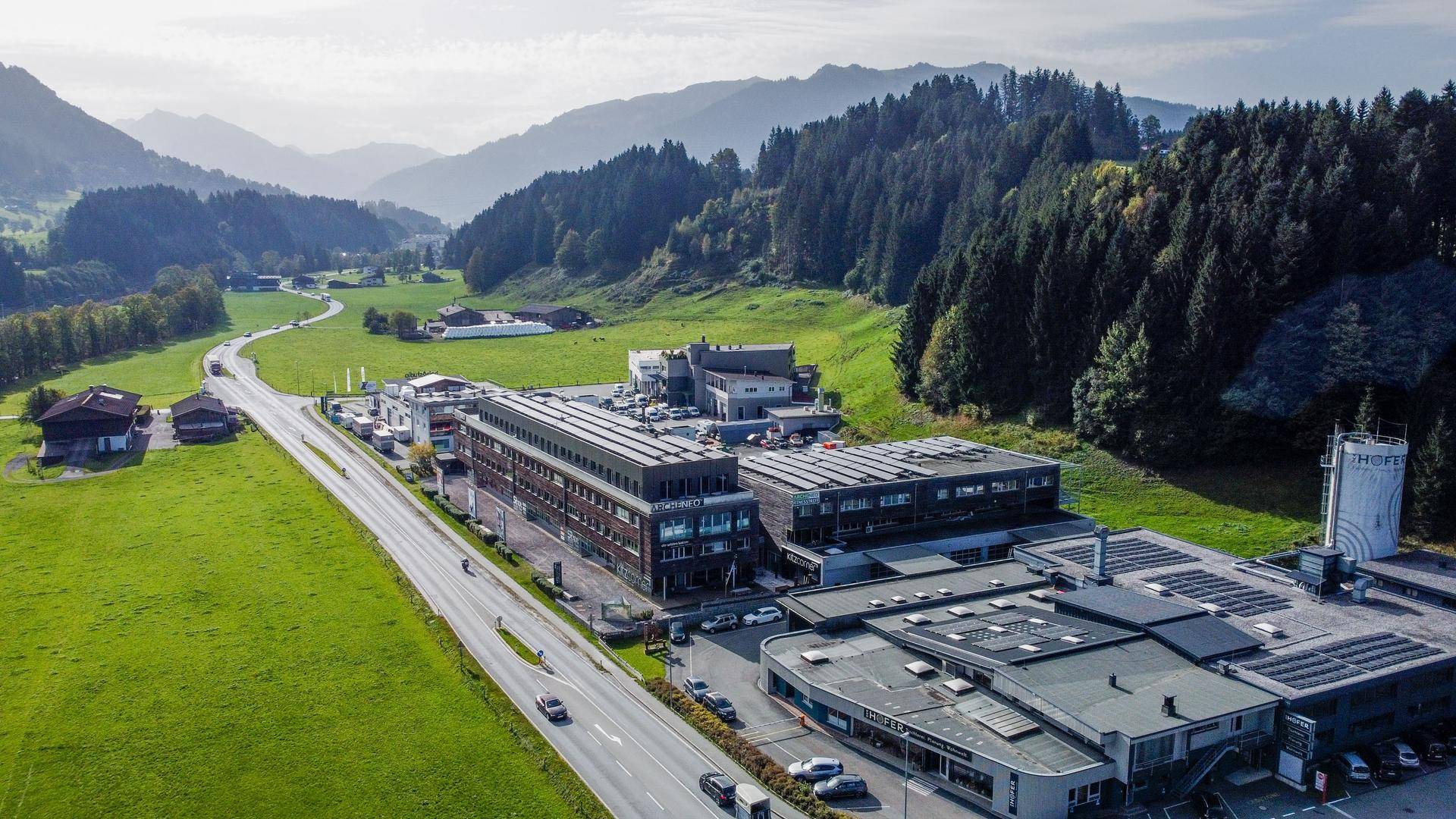 Luftaufnahme eines Gewerbegebiets mit mehreren Gebäuden, Parkplätzen und einer Straße in bergiger Landschaft.