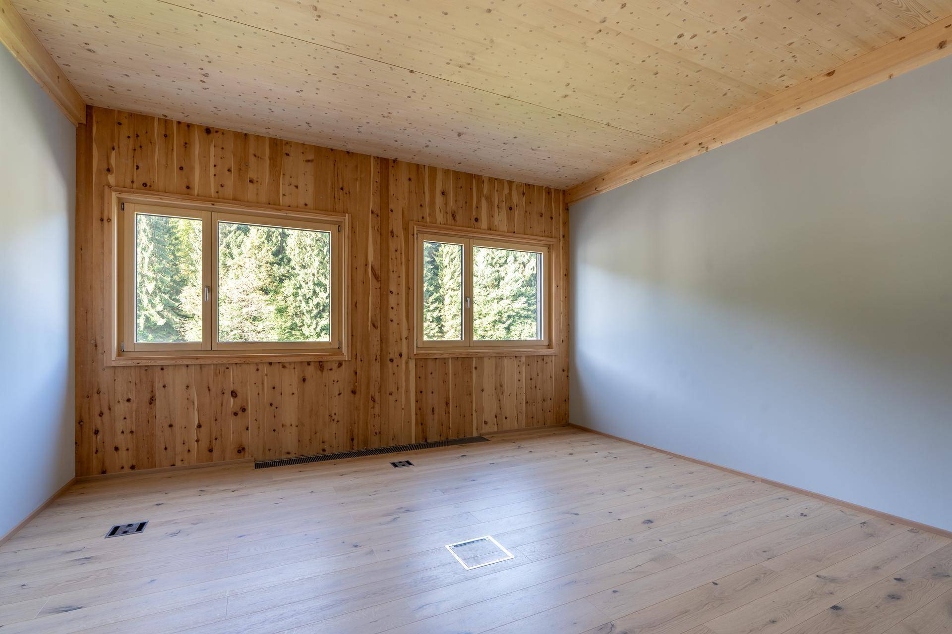Heller Raum mit Holzwand und zwei Fenstern, die einen Blick auf grüne Bäume bieten.