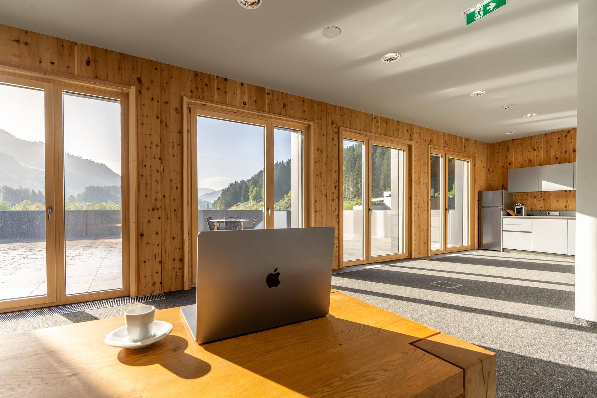 Heller Büroraum mit Holzwänden, großen Fenstern, Zugang zur Terrasse und einer kleinen Küchenzeile.