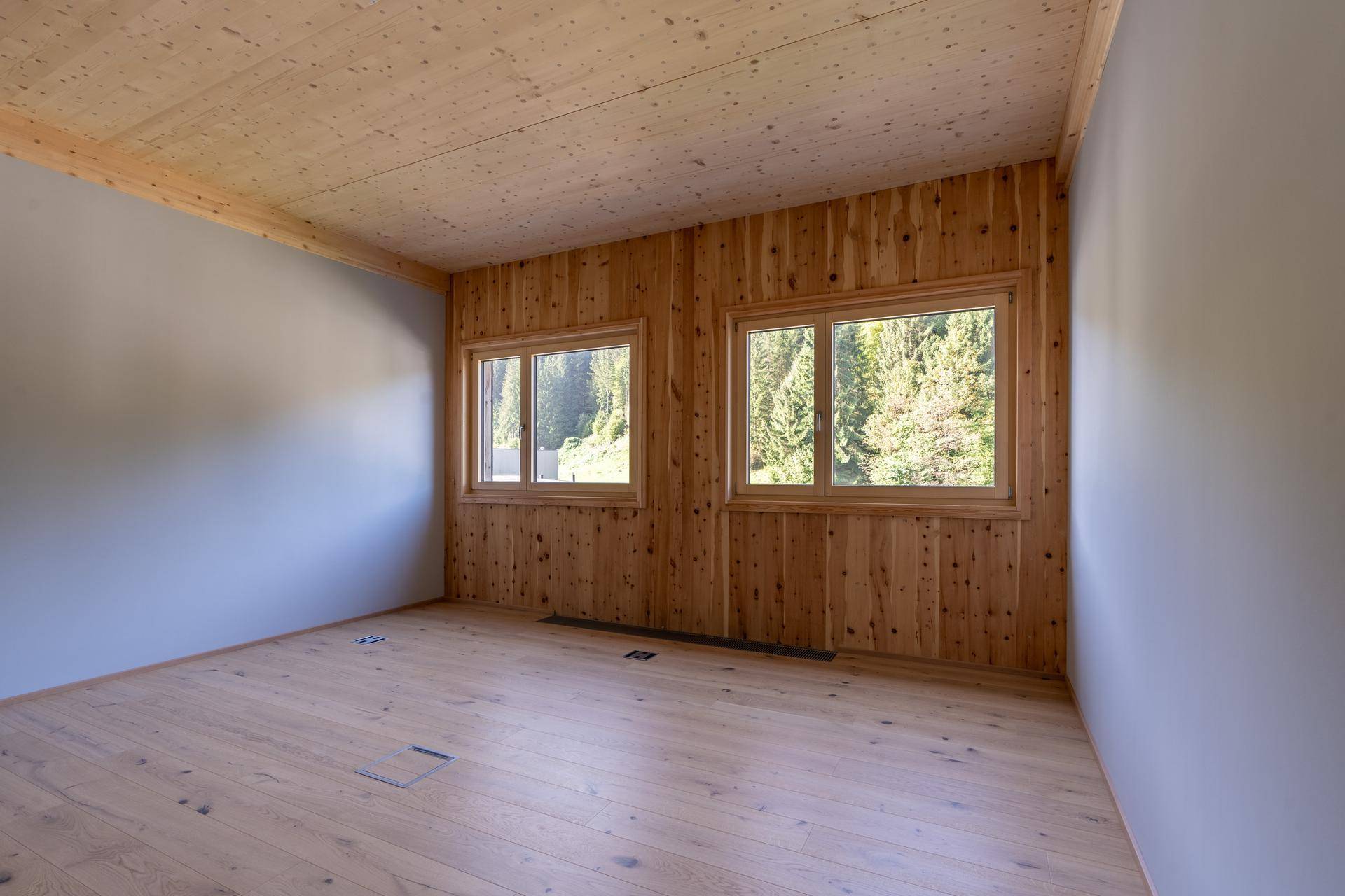 Heller Raum mit Holzwand und zwei Fenstern, die einen Blick auf grüne Bäume bieten.