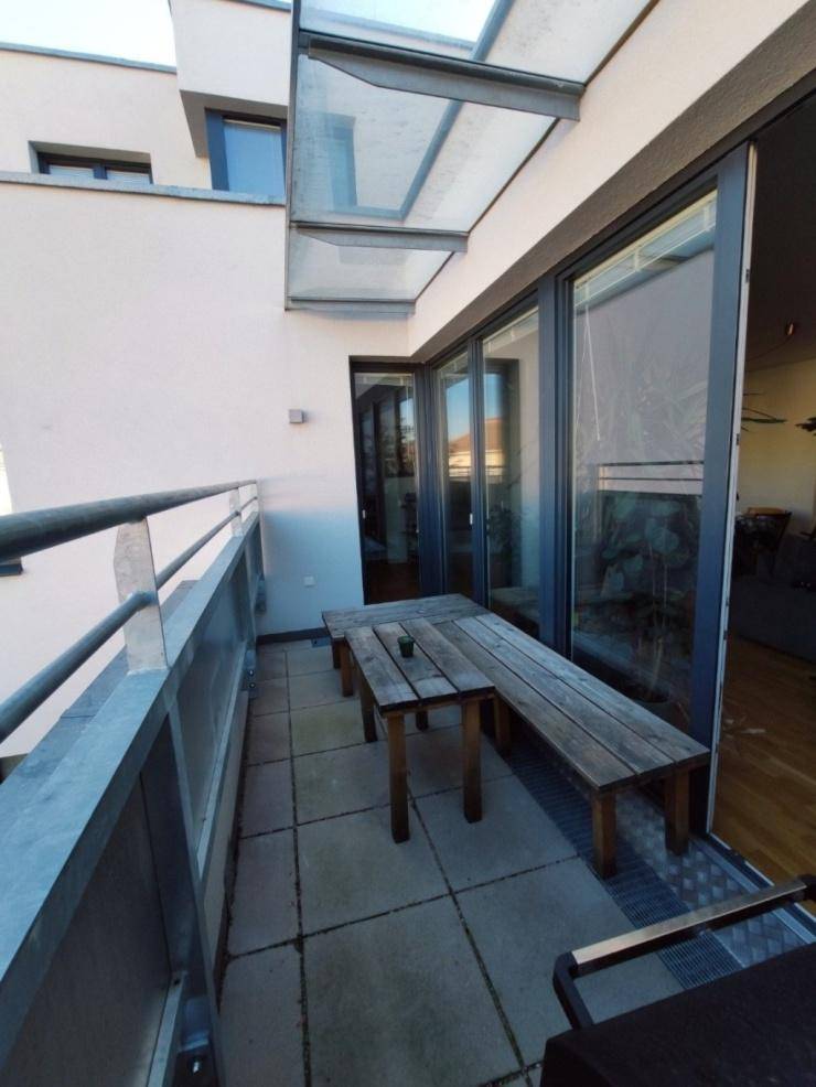Schmaler Balkon mit Holztisch und Bank, überdacht mit einer Glasüberdachung, bietet einen Außenbereich.