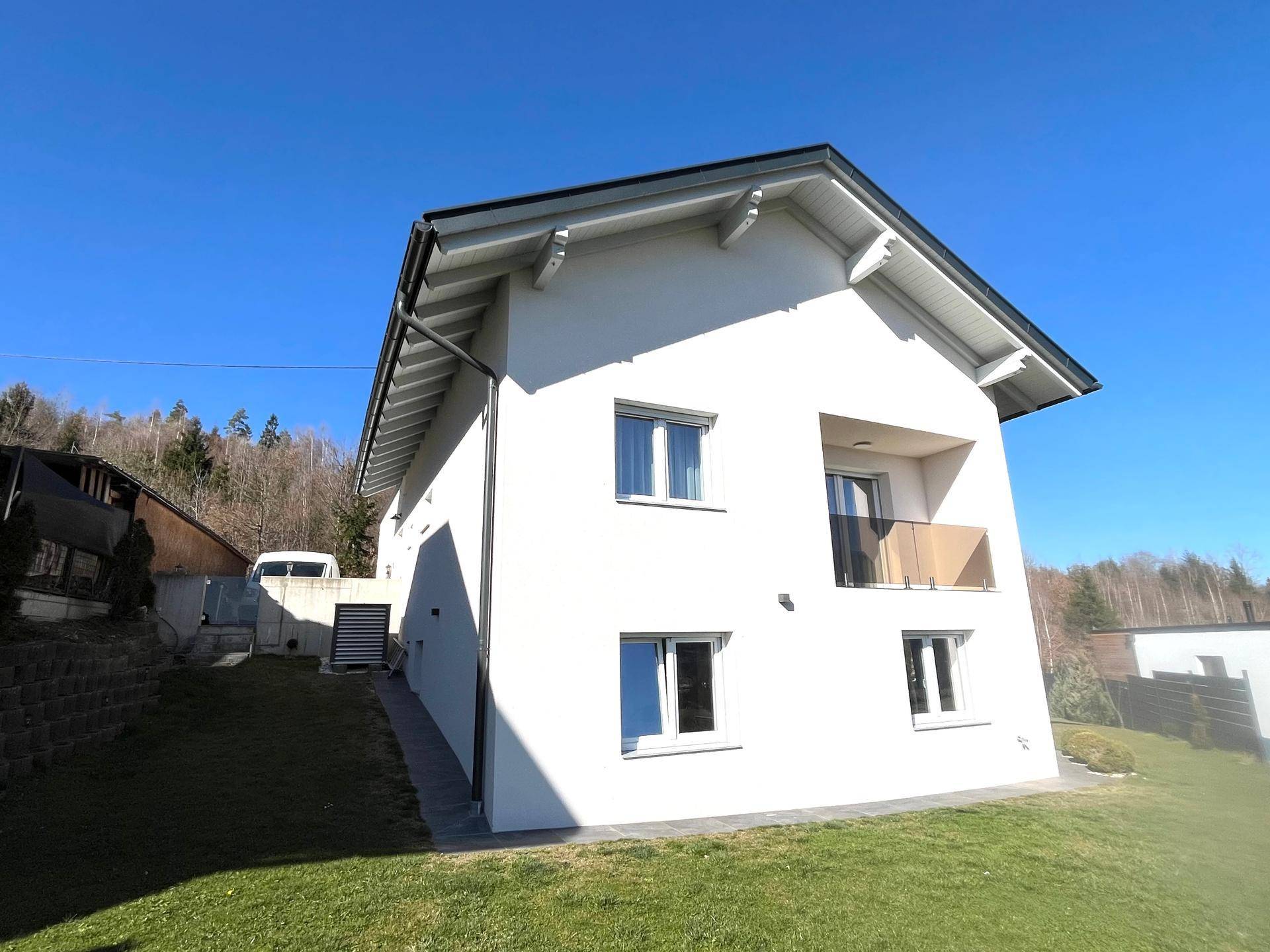 Modernes Einfamilienhaus mit weißer Fassade, Satteldach und kleinem Balkon, umgeben von Grünflächen.