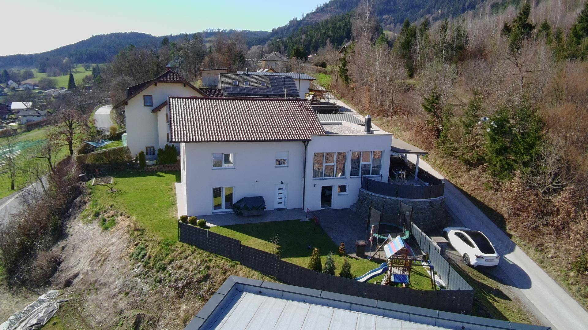 Luftaufnahme eines modernen Hauses mit Flachdach-Anbau, Garten und Spielplatz in ländlicher Umgebung.