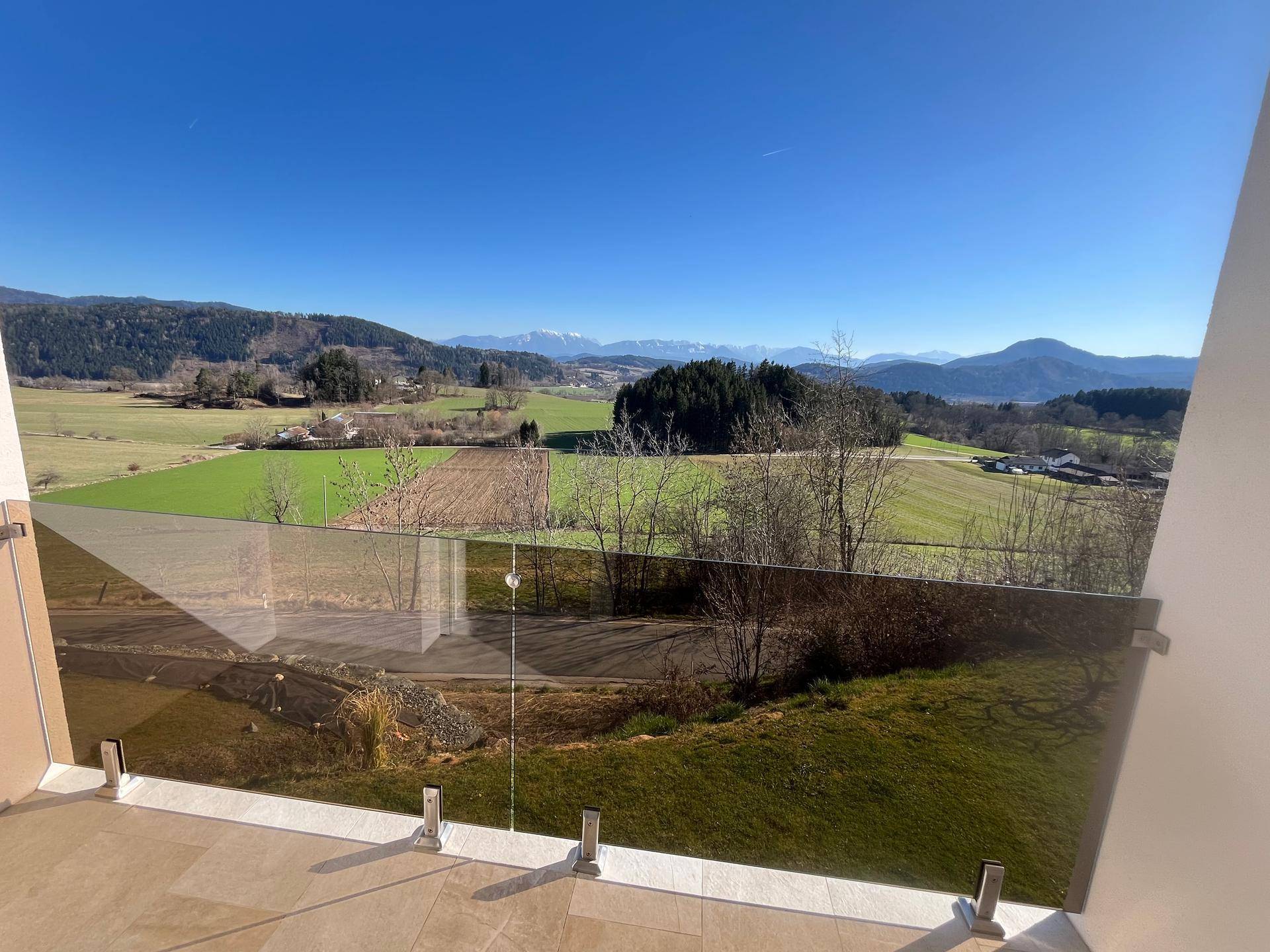 Balkon mit Glasgeländer und weitem Blick über grüne Felder und schneebedeckte Berge.
