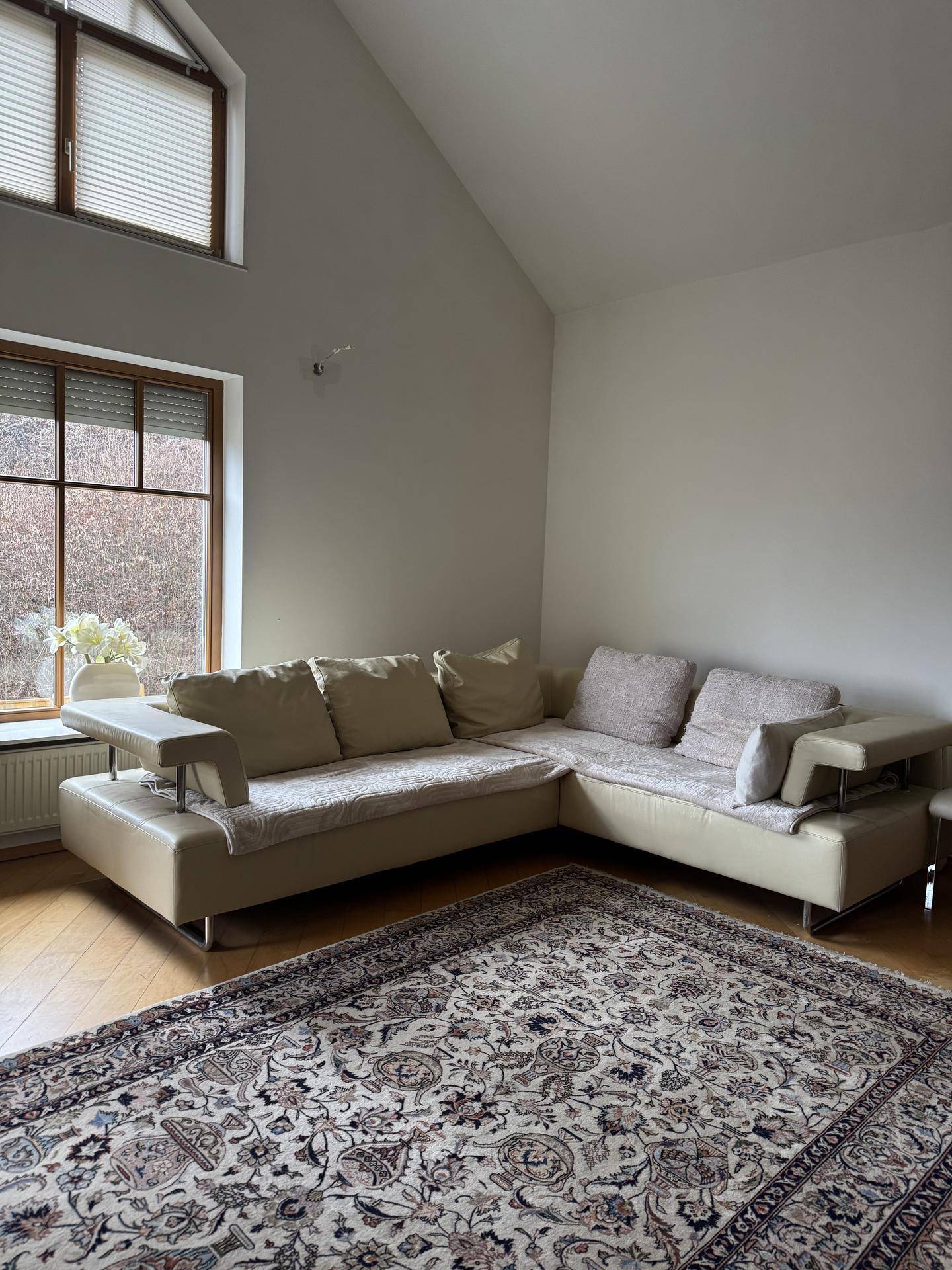 Helles Wohnzimmer mit hoher Decke, beigem Ecksofa und gemustertem Teppichboden.