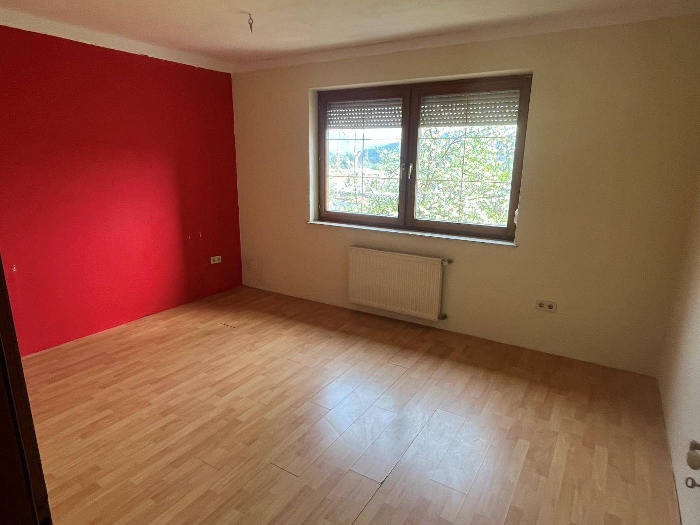 Schlafzimmer mit roter Akzentwand, Parkettboden und großem Fenster.