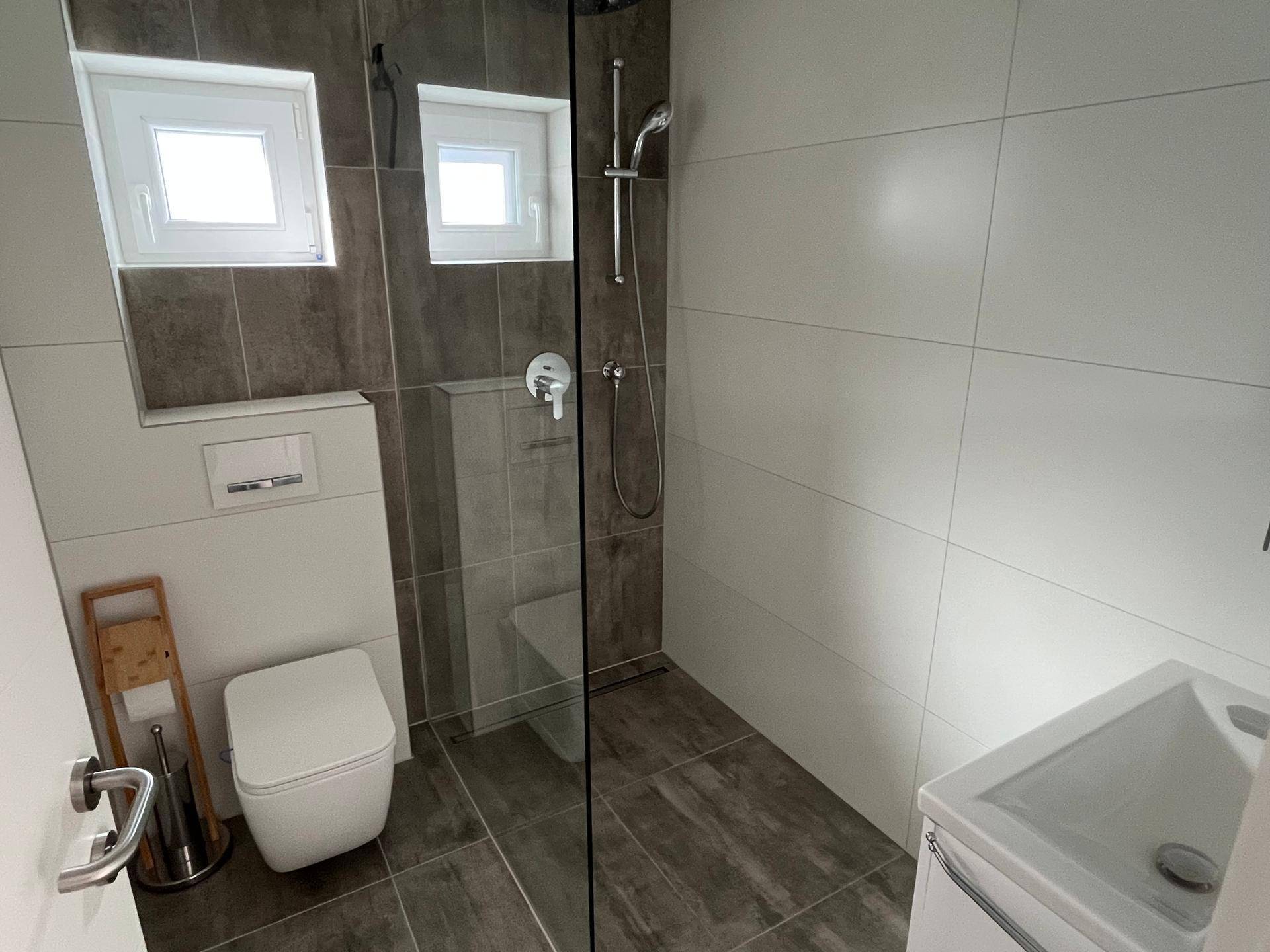 Modernes Duschbad mit bodengleicher Dusche, Wand-WC und zwei Fenstern für Tageslicht.
