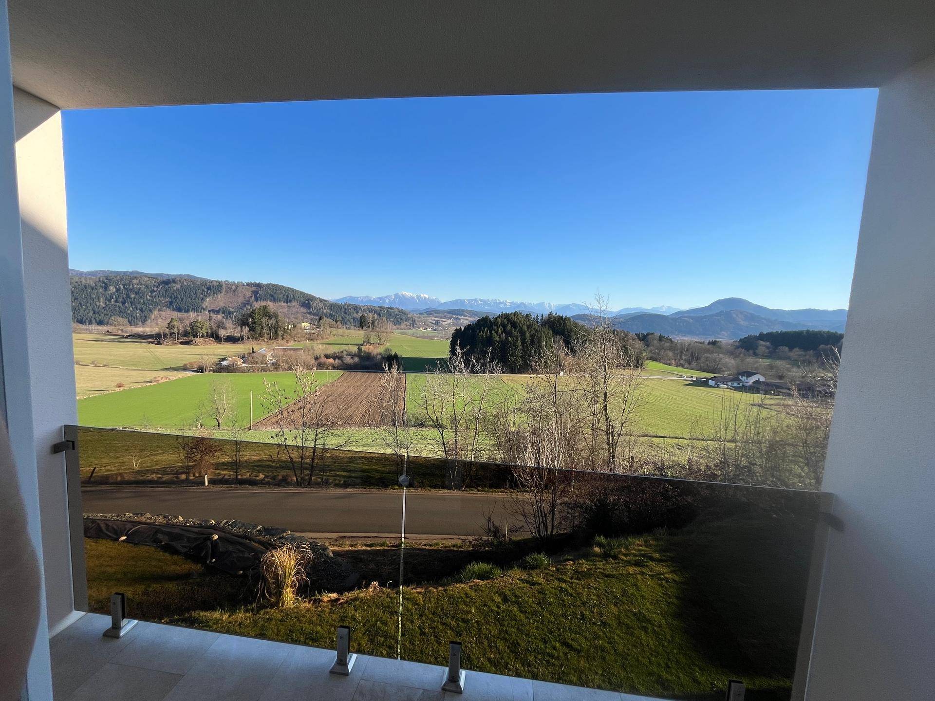 Weitläufiger Blick vom Balkon über grüne Felder und ferne Berge unter blauem Himmel.